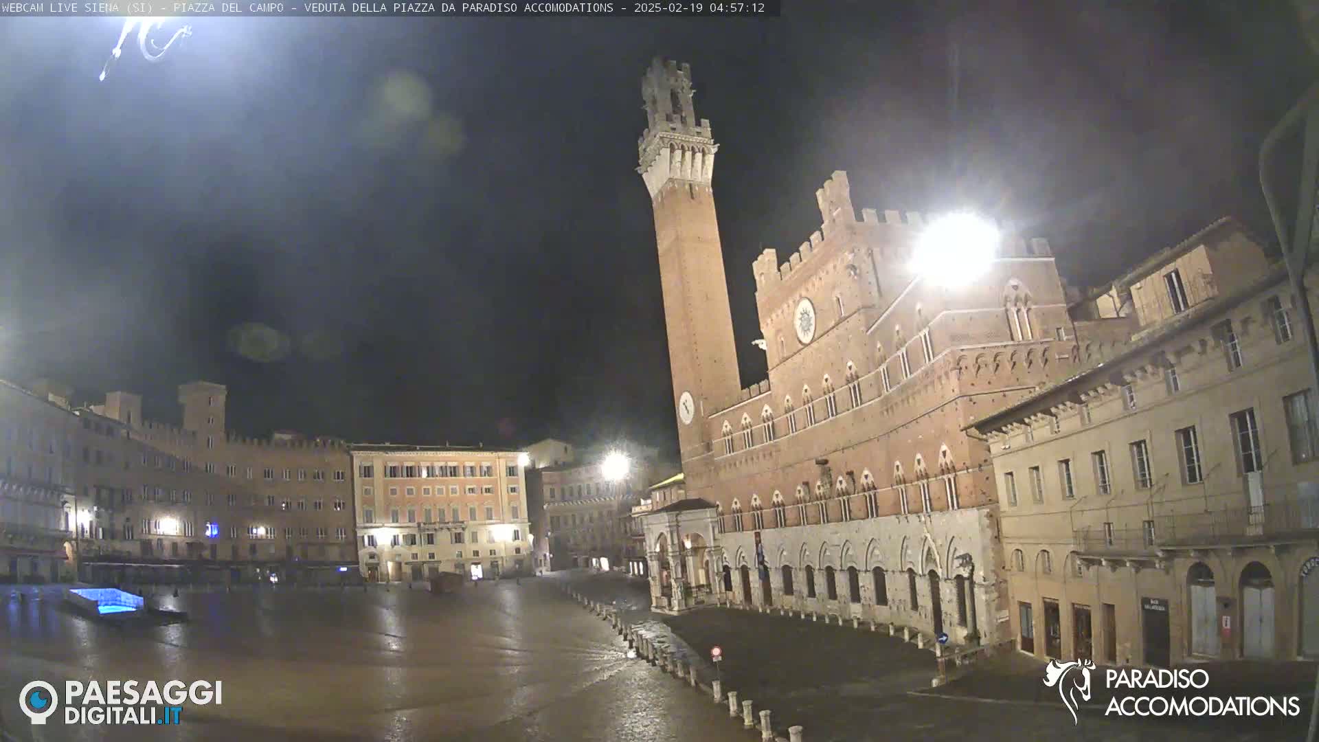 A nighttime view of a large, empty square in a European city, with a tall brick tower and surrounding buildings illuminated under a dark, possibly rainy sky.
