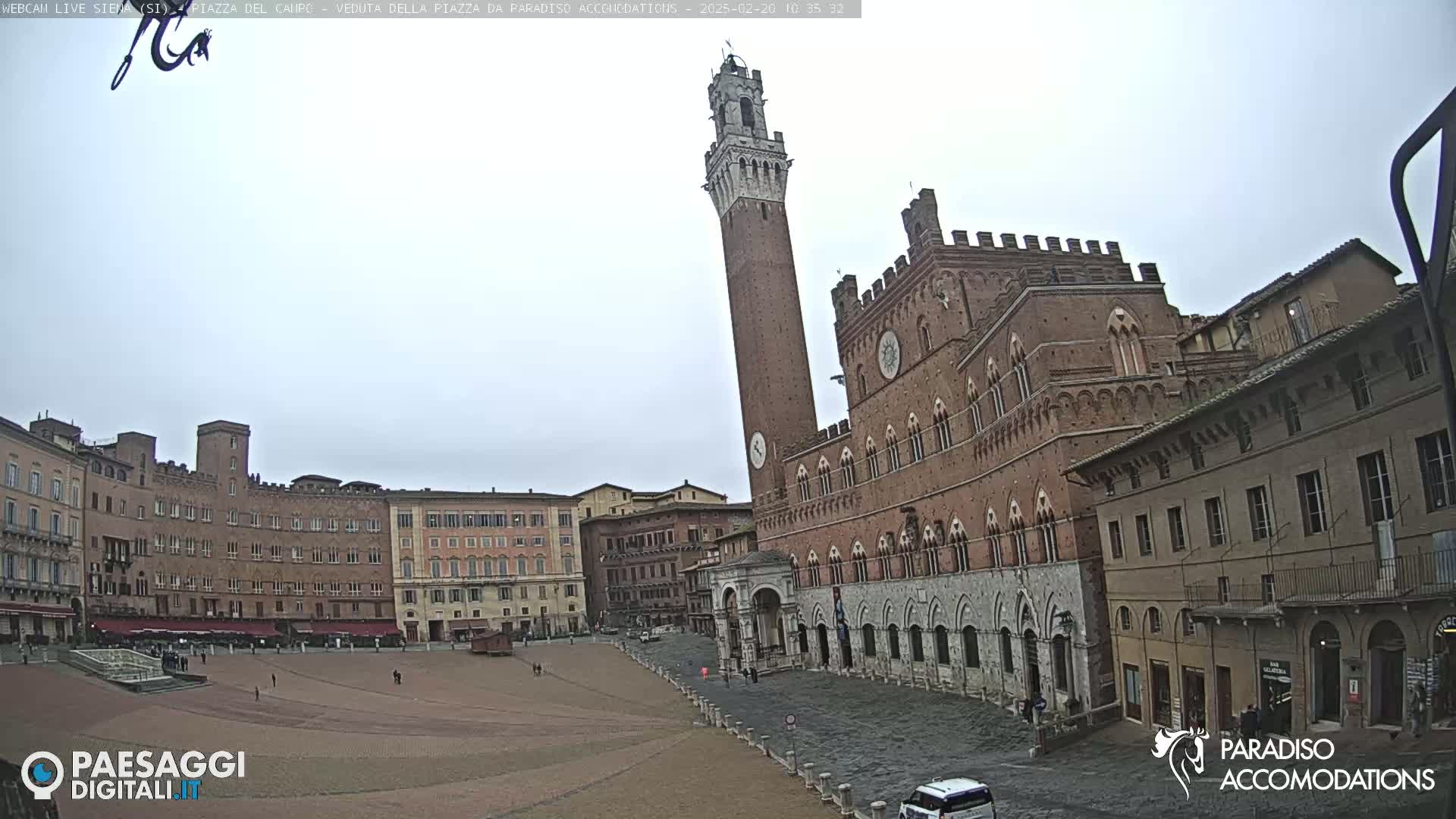 A large, brick plaza in a city, under an overcast sky, is surrounded by medieval-style buildings, including a tall tower.