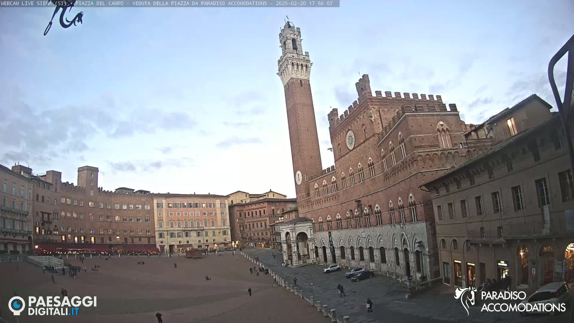 A large, brick piazza in Italy is mostly empty except for a few pedestrians, with a tall, slender tower and a large, ornate building dominating the scene under a mostly clear, dusky sky.