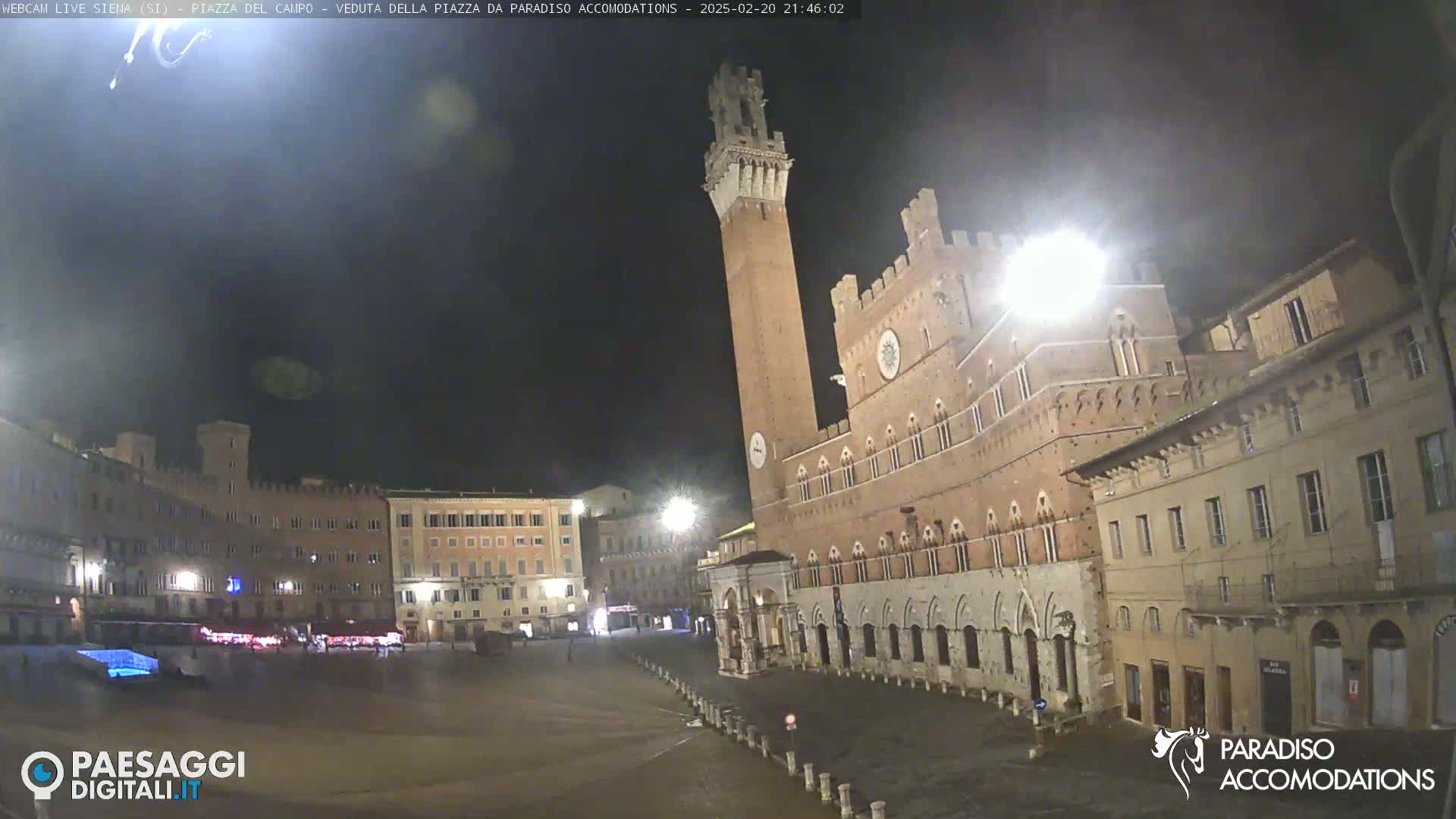 A nighttime view of a large, empty plaza in a European city, featuring an imposing brick building with a tall tower, under a dark sky.