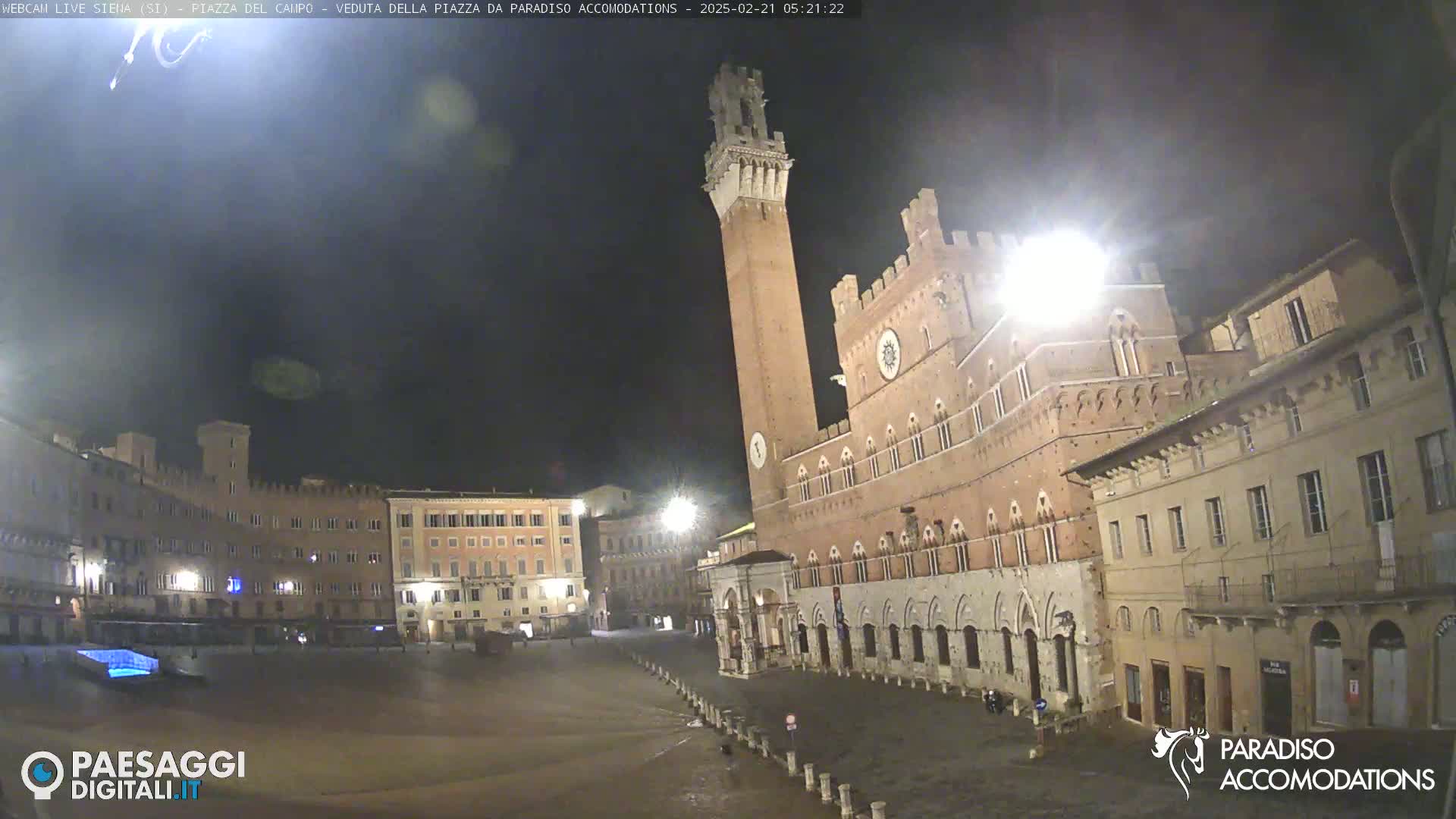 A nighttime view of a large, mostly empty plaza with historic brick buildings, including a tall tower, under a dark sky.