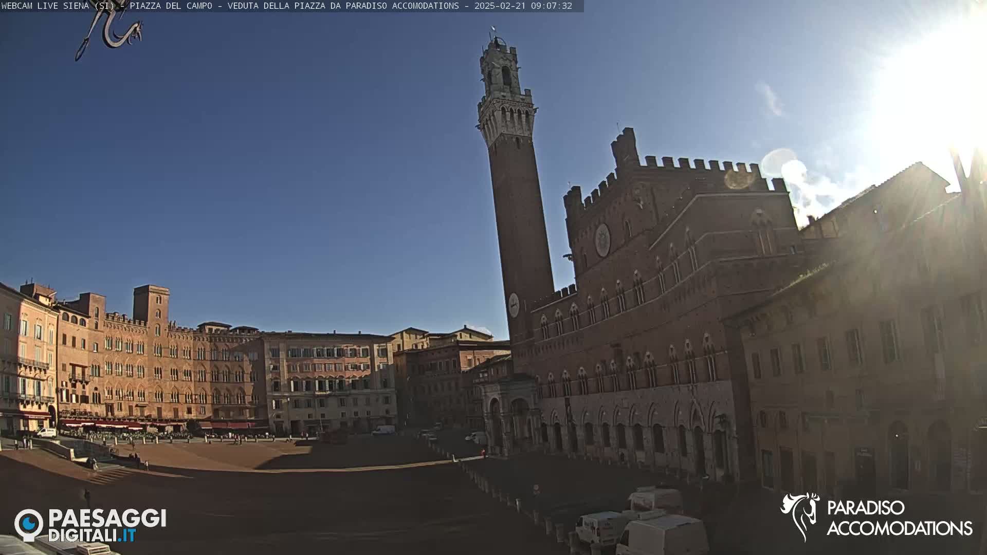 A sunny day in a large, mostly empty piazza surrounded by historic brick buildings, including a tall tower and a large palace.