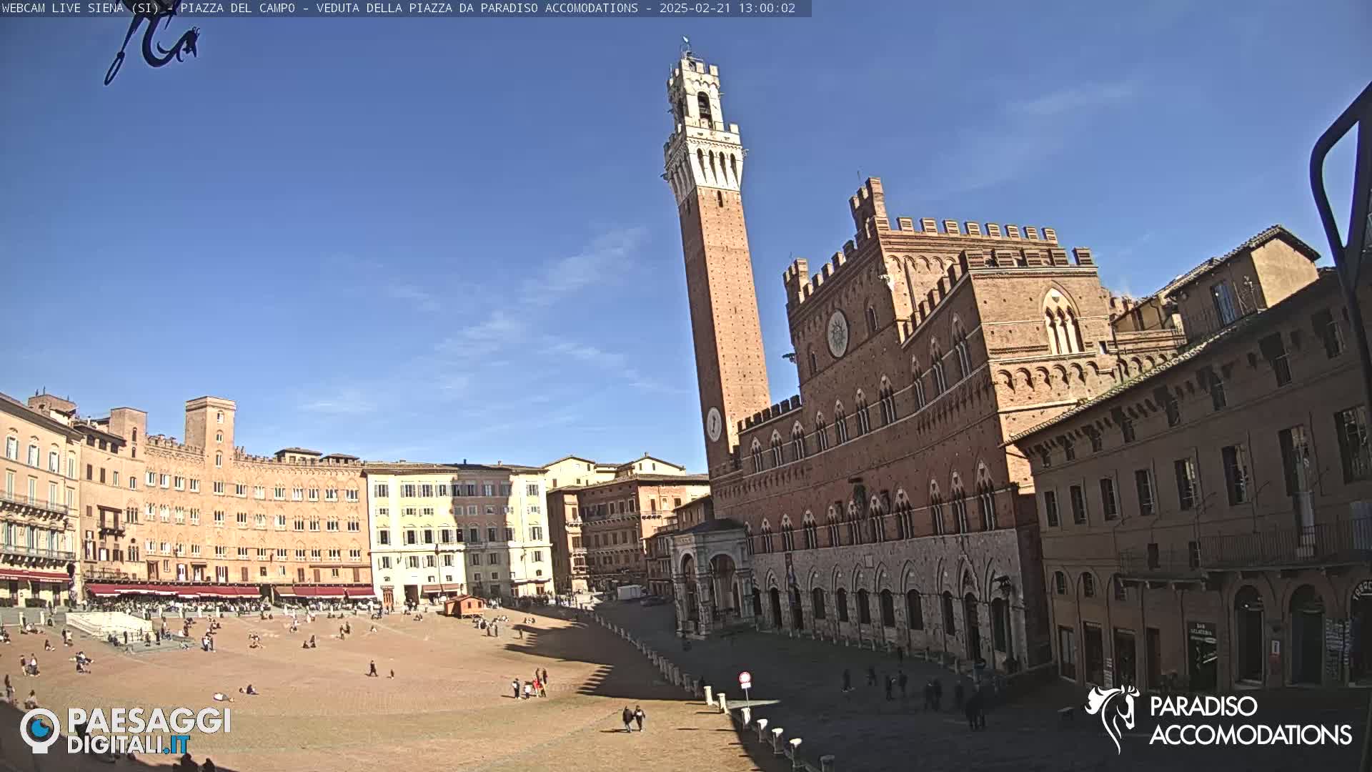 A large, sunlit brick piazza in Italy, featuring a tall tower and surrounding historical buildings under a clear blue sky.