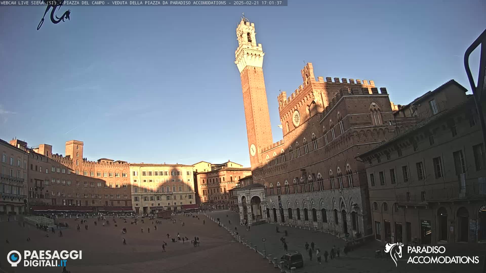 A large, brick public square in the late afternoon sun, features a tall, slender tower and a large, ornate building.