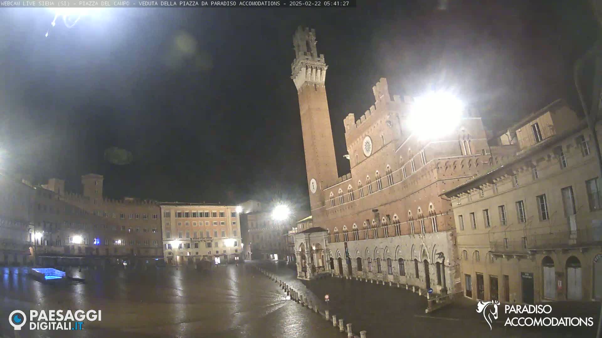 A large, empty, wet piazza at night, surrounded by historic brick buildings, one of which features a tall tower.