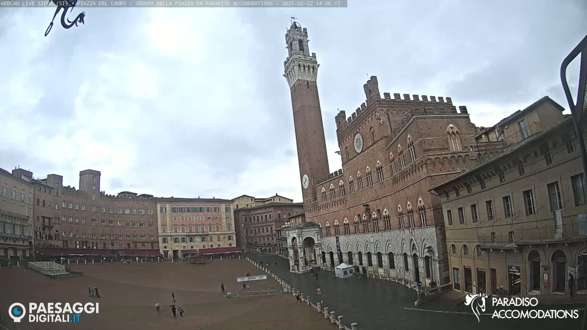 A large, empty plaza in a city with red-brick buildings, including a tall tower and a palace, under an overcast sky.