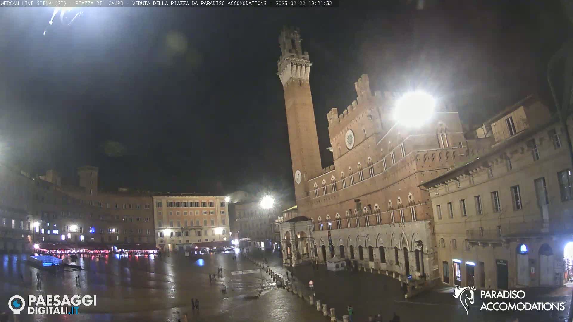A rain-slicked, nighttime view of a large, brick public square in Italy, dominated by a tall, slender tower and a grand, medieval-style building.