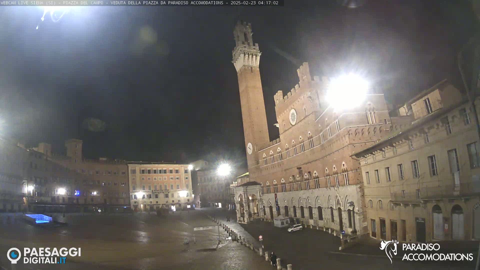 A nighttime, mostly clear view of a large, empty plaza in a European city, centered on a tall, brick tower and adjacent building.