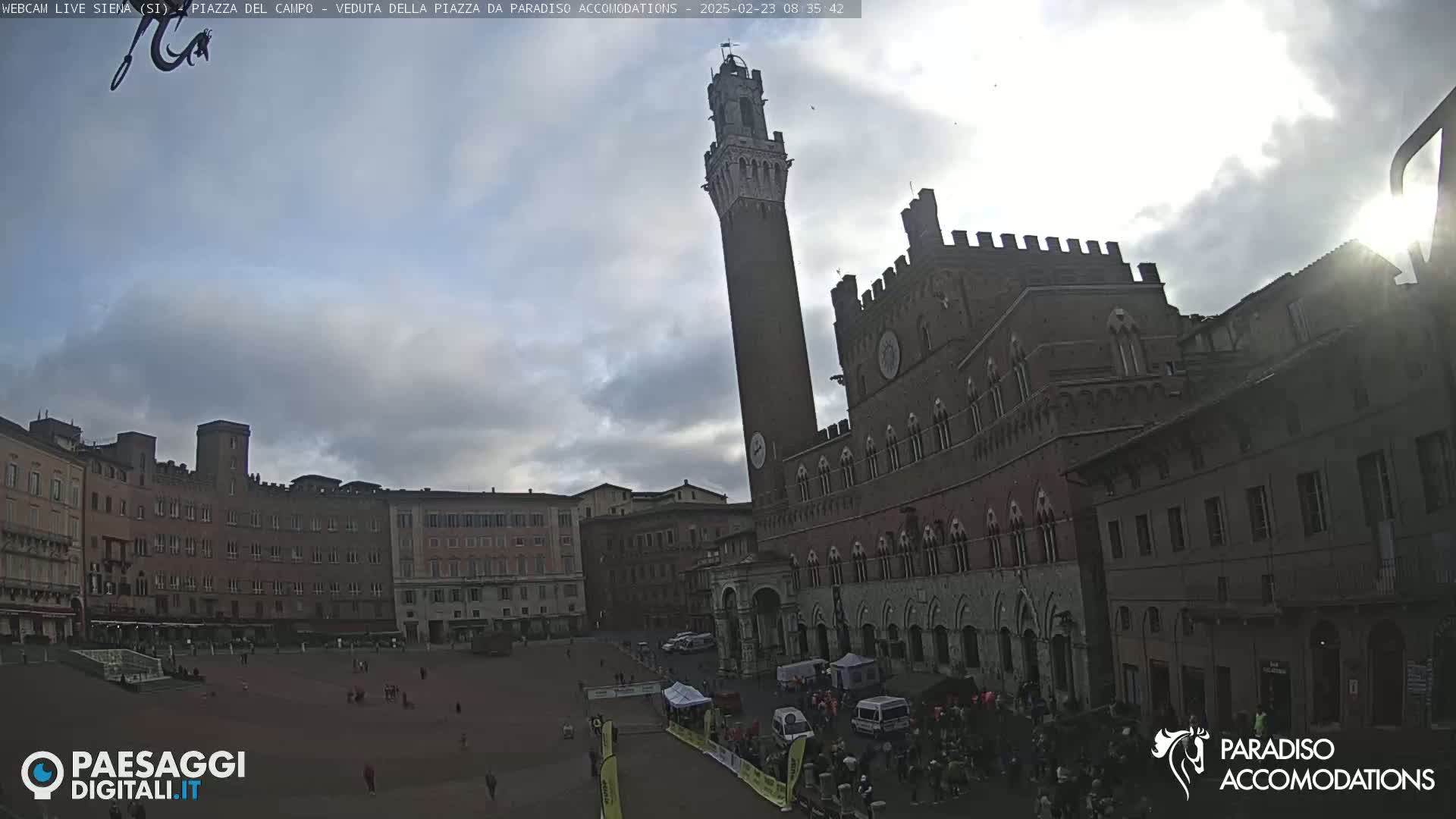 A large, mostly empty plaza in front of a tall, red brick tower and palazzo under a partly cloudy sky.