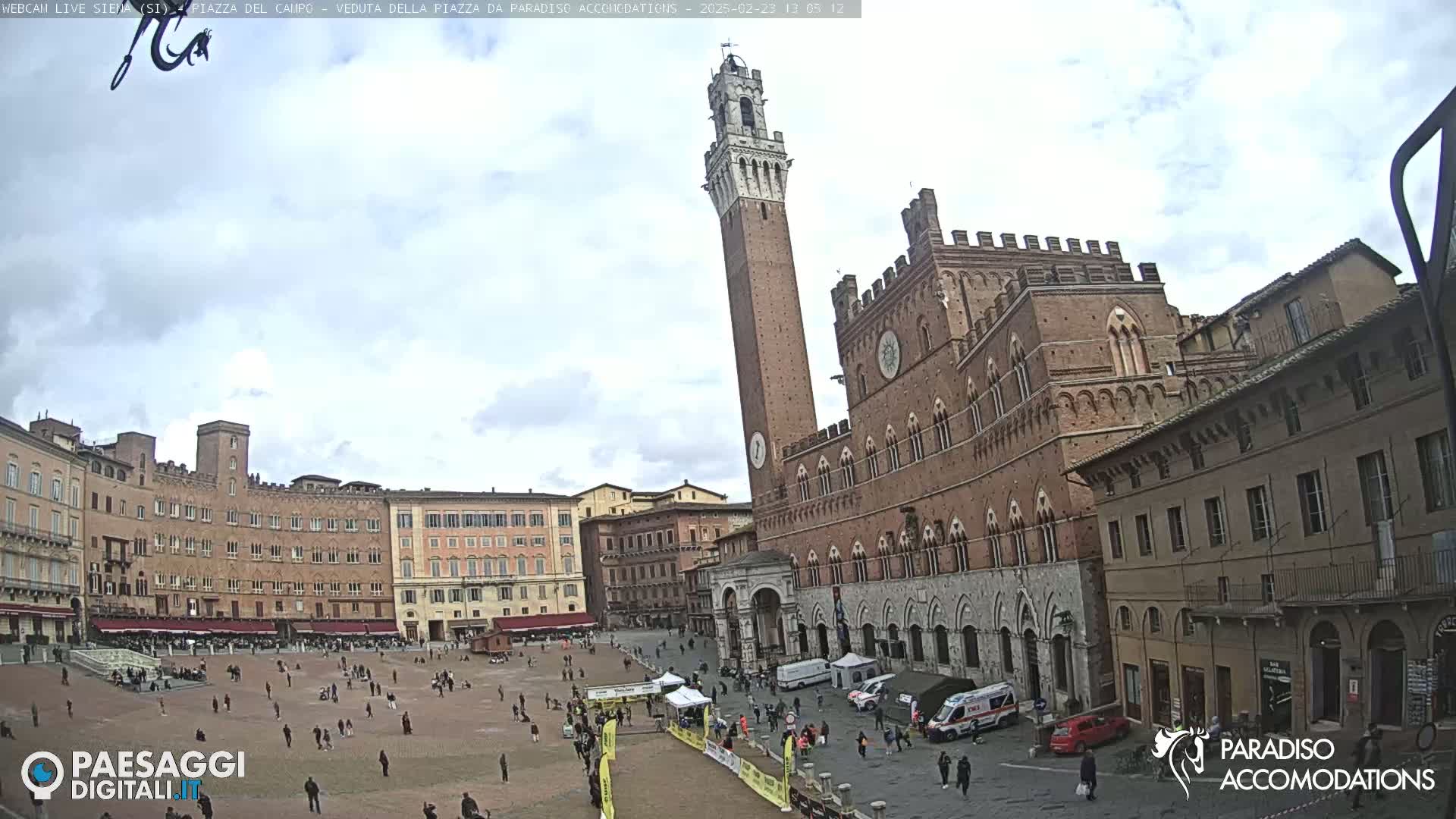 A large, brick piazza in Italy is mostly empty except for a few people scattered around, with a tall brick tower and several multi-story buildings surrounding it under a mostly cloudy sky.