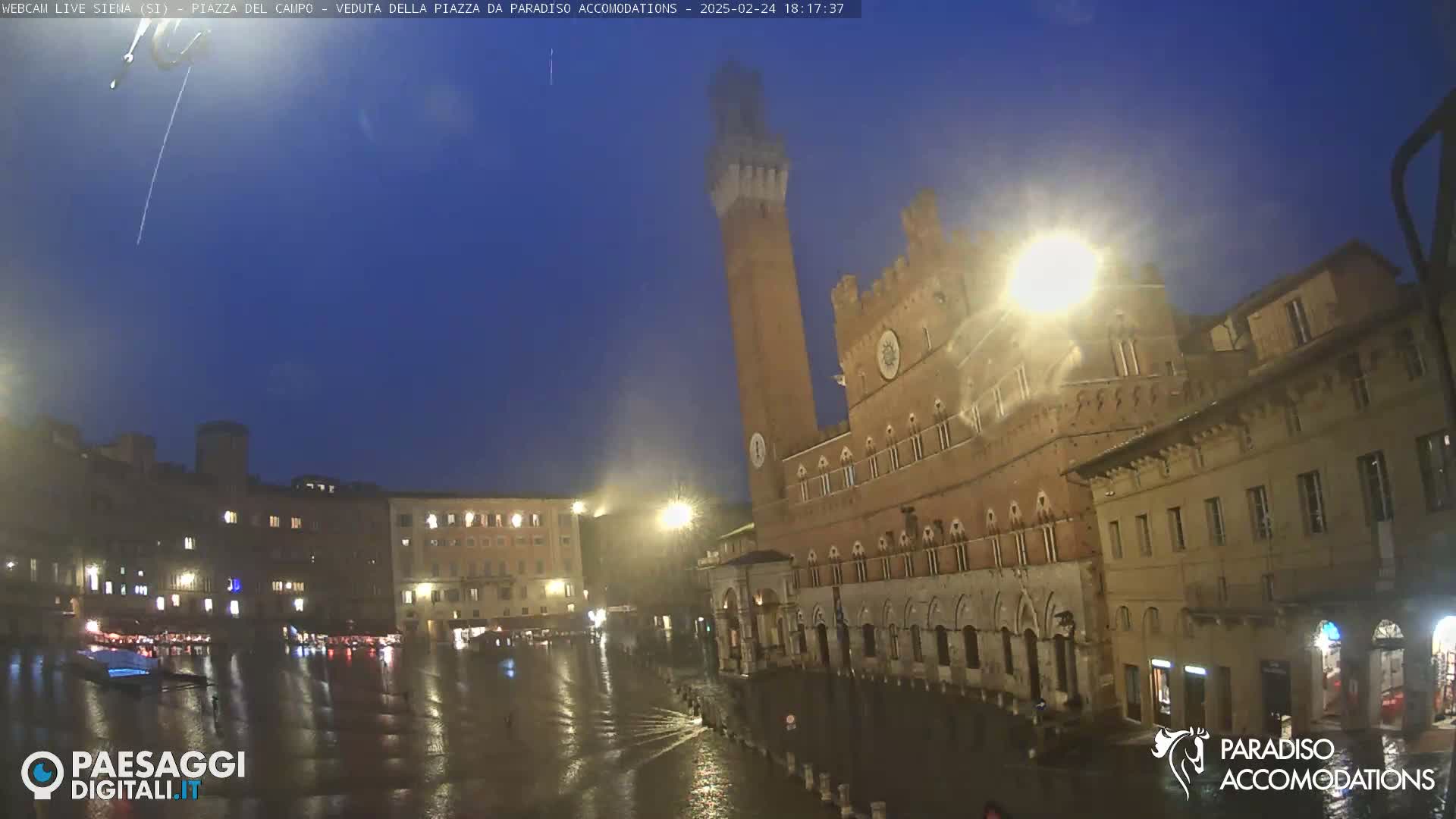 A rain-soaked piazza in an Italian city at twilight, featuring a tall brick tower and large stone buildings illuminated by streetlights.