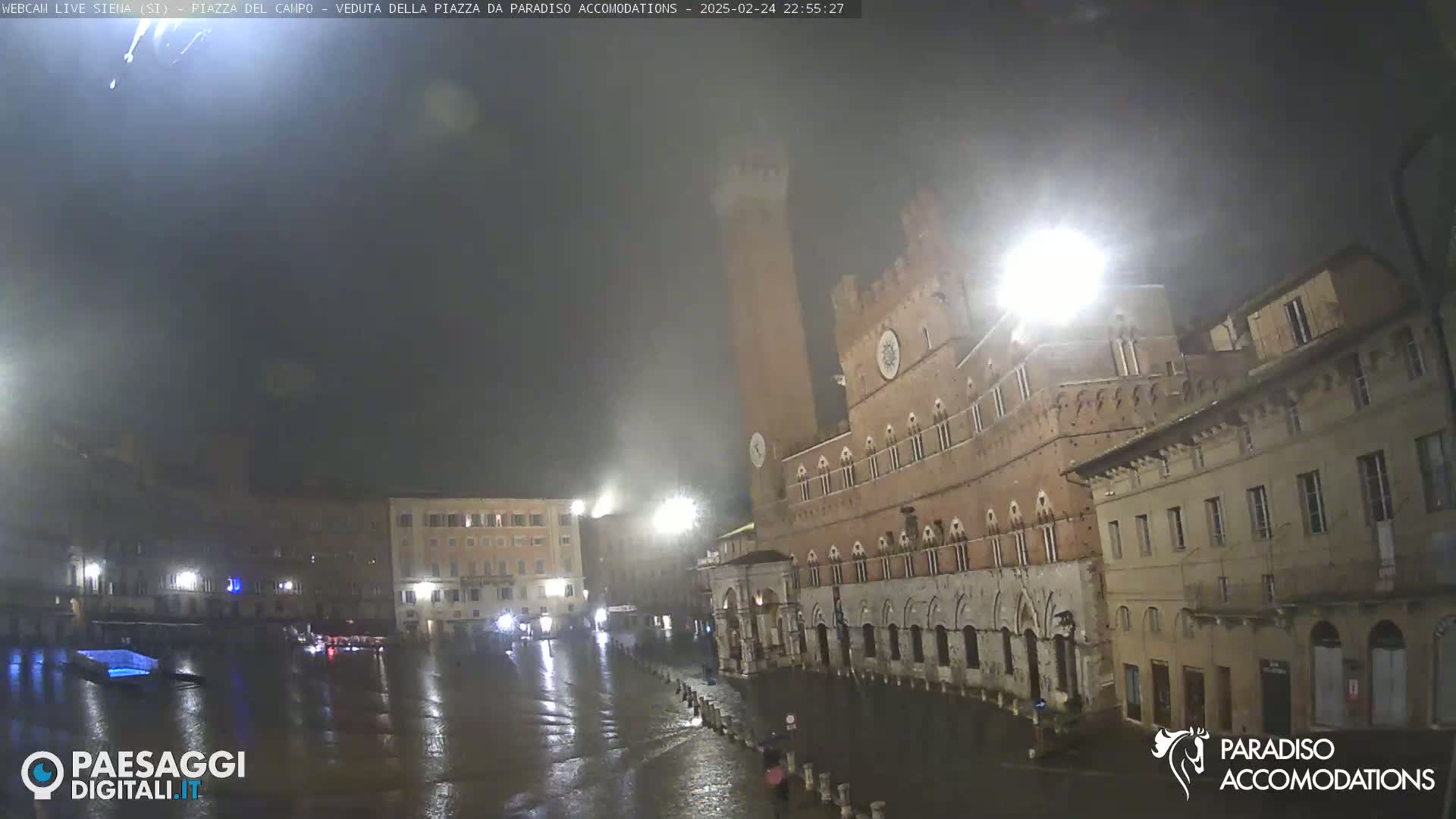 A rain-soaked town square at night, illuminated by streetlights, features a tall brick tower and surrounding buildings.