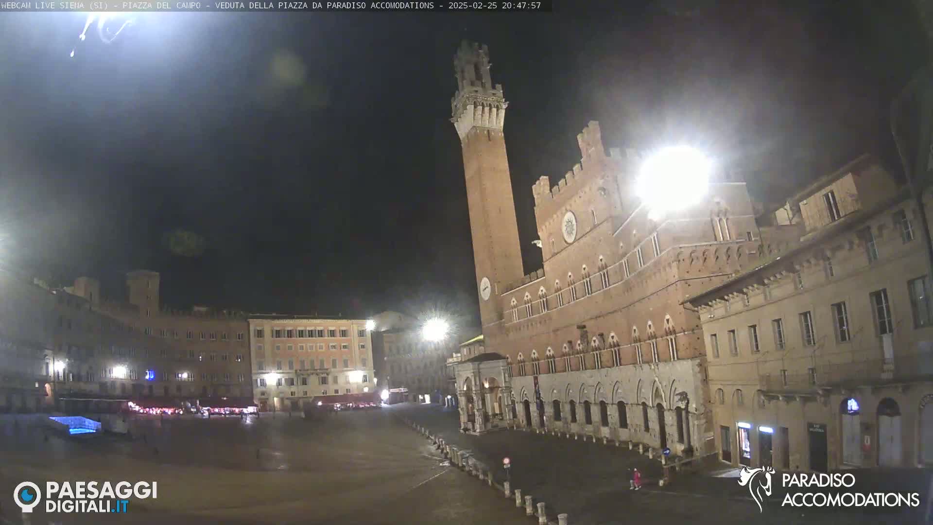 A large, empty piazza at night, illuminated by streetlights, is surrounded by historic brick buildings, including a tall tower.