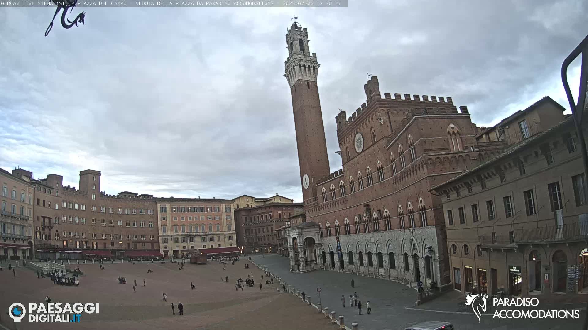 A large, brick public square in Italy, under a cloudy sky, features a tall, slender tower and multiple historic buildings.
