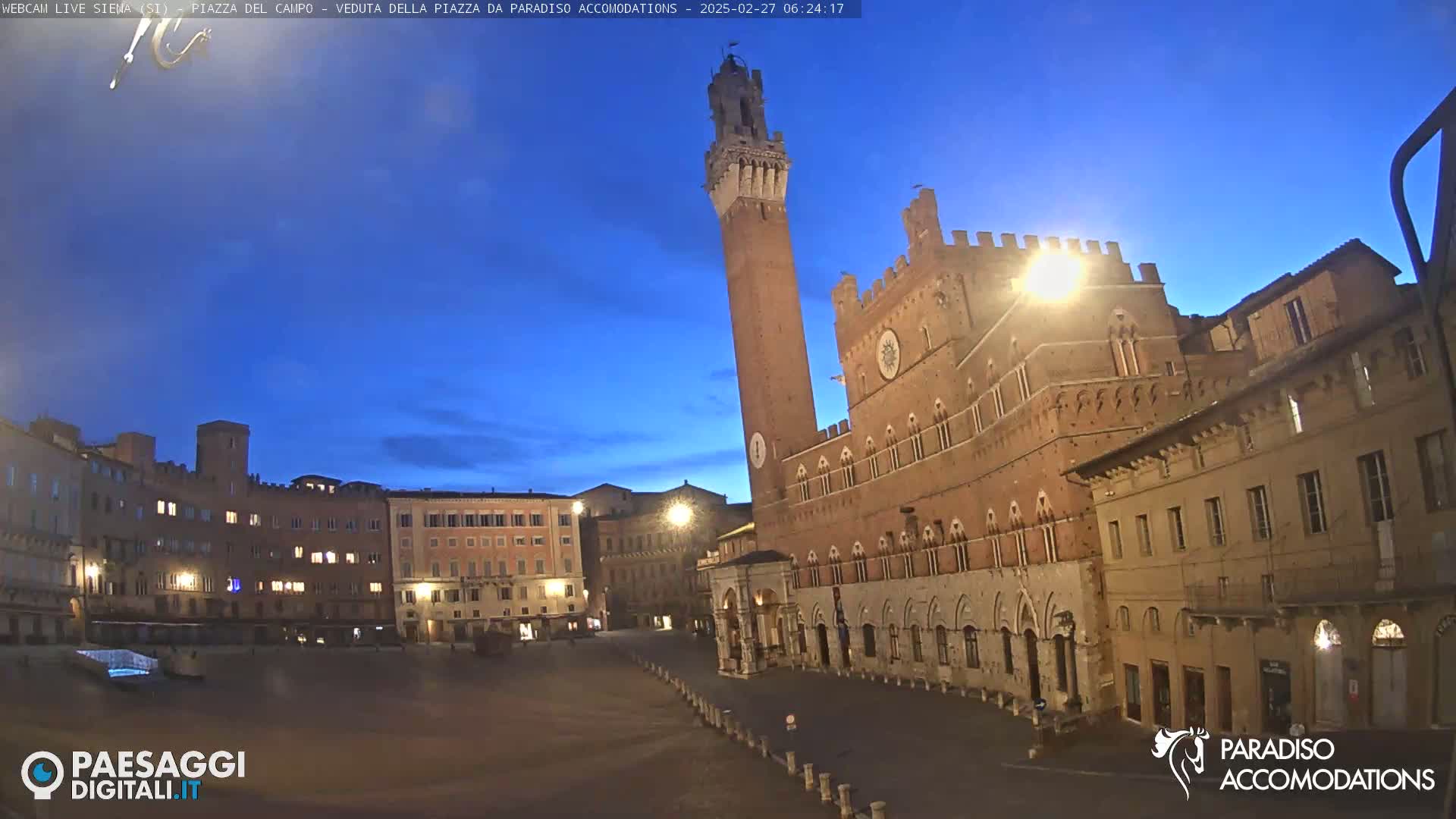 A large, empty town square at twilight is surrounded by medieval-style buildings, including a tall brick tower.