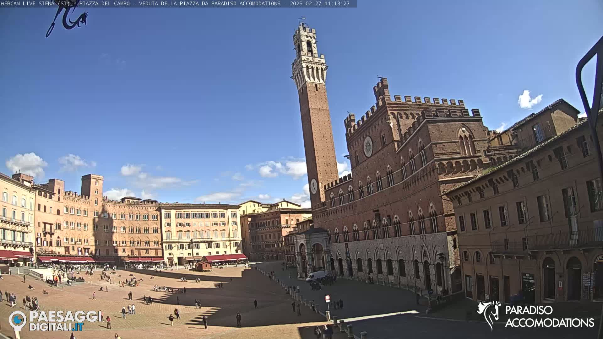 A large, brick piazza in bright sunlight is surrounded by historic buildings, including a tall, slender tower and a grand palace.