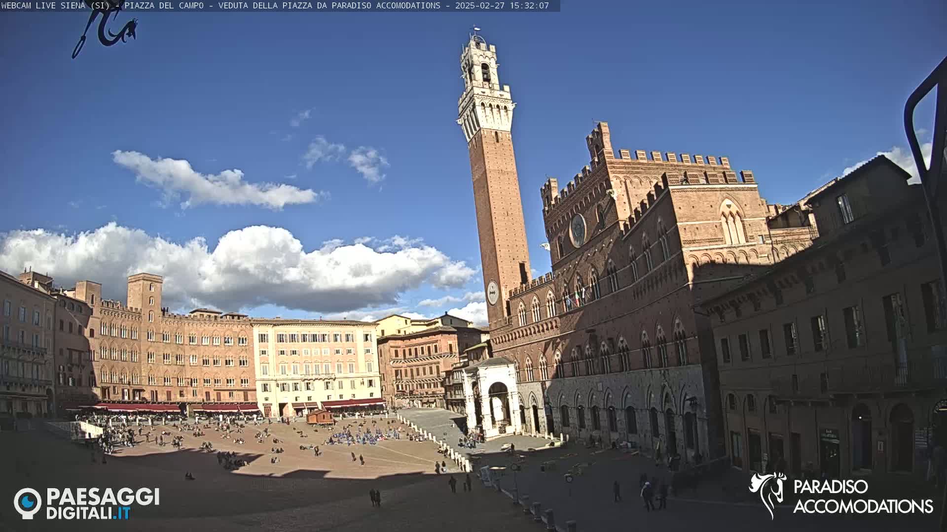 A sunny day in a large, brick-colored plaza, featuring several historical buildings, including a tall tower and a large palace, with people sparsely scattered throughout.