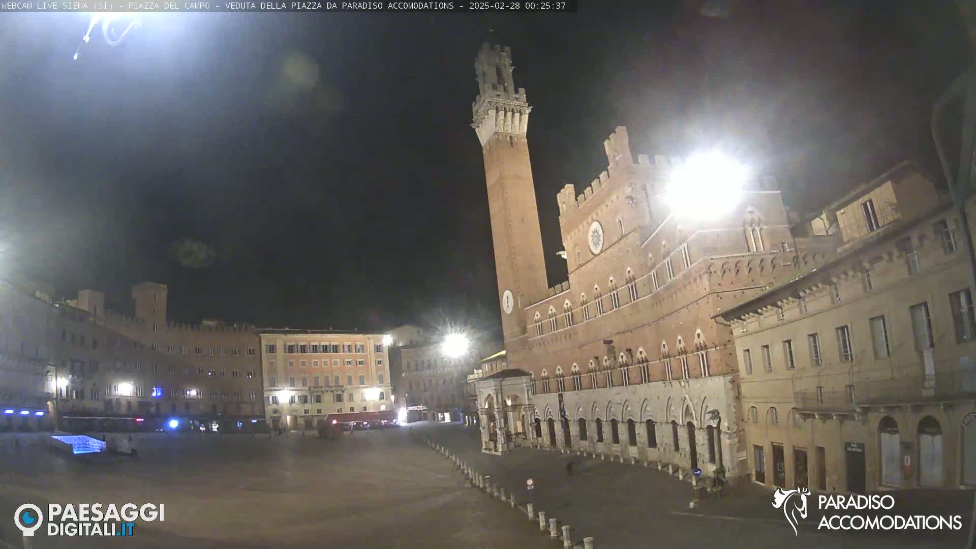 A mostly empty, brightly lit plaza at night in a European city features a large brick building with a tall tower, surrounded by other buildings.