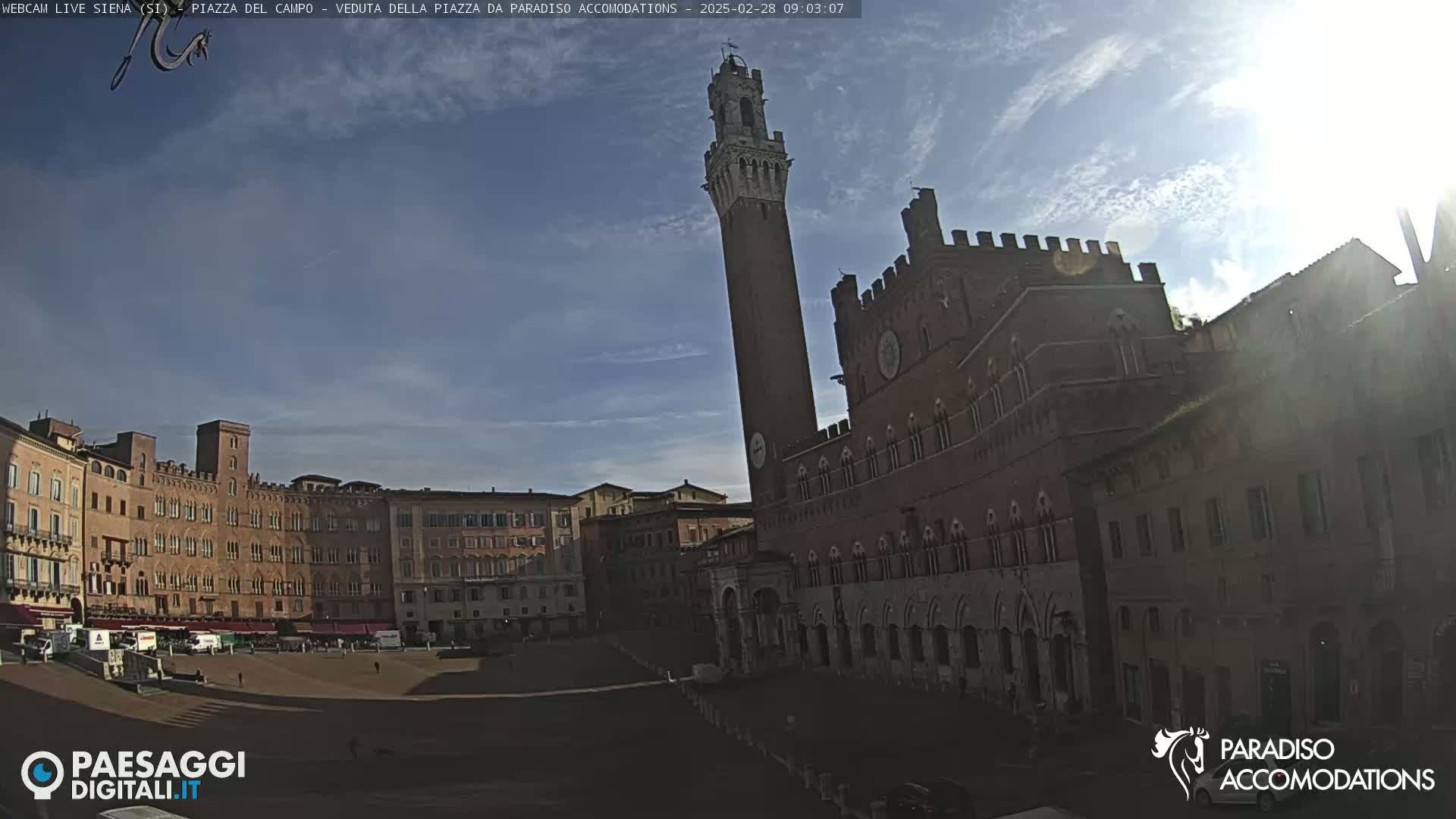 A large, sunlit piazza in Siena, Italy, is surrounded by historic brick buildings, including a tall tower and a grand palazzo.