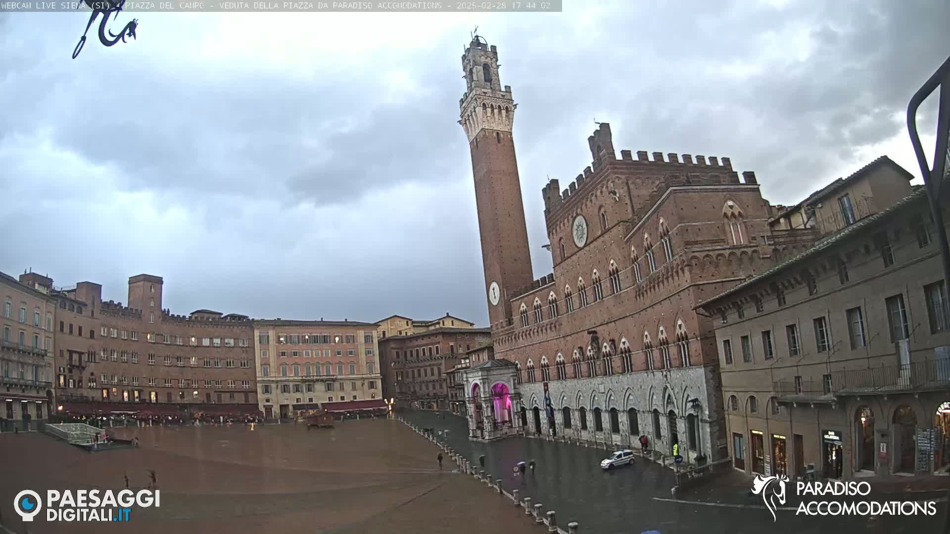 A large, brick piazza in a city is seen on an overcast day with some wet pavement; a tall, slender tower stands prominently next to a large brick building.