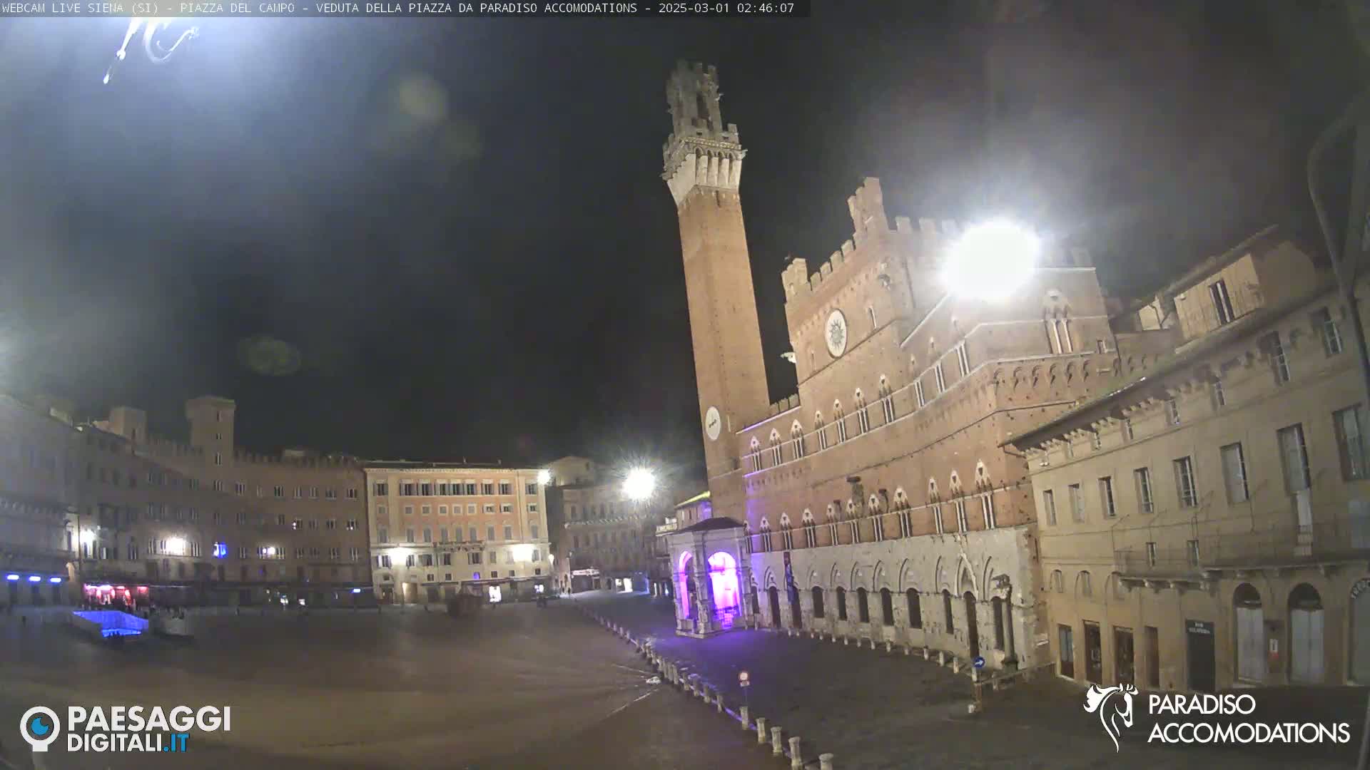 A mostly empty, large town square at night, featuring a tall brick tower and surrounding medieval-style buildings under a dark sky.
