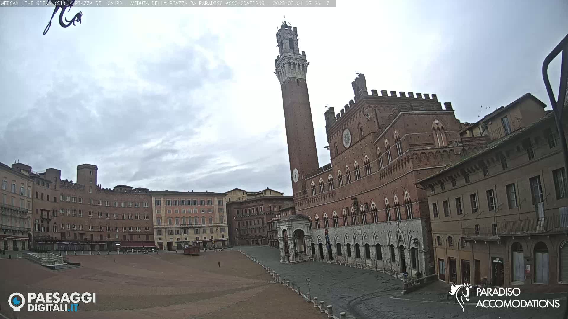 A large, brick public square in an Italian city, under an overcast sky, features a tall, slender tower and a grand, ornate building.