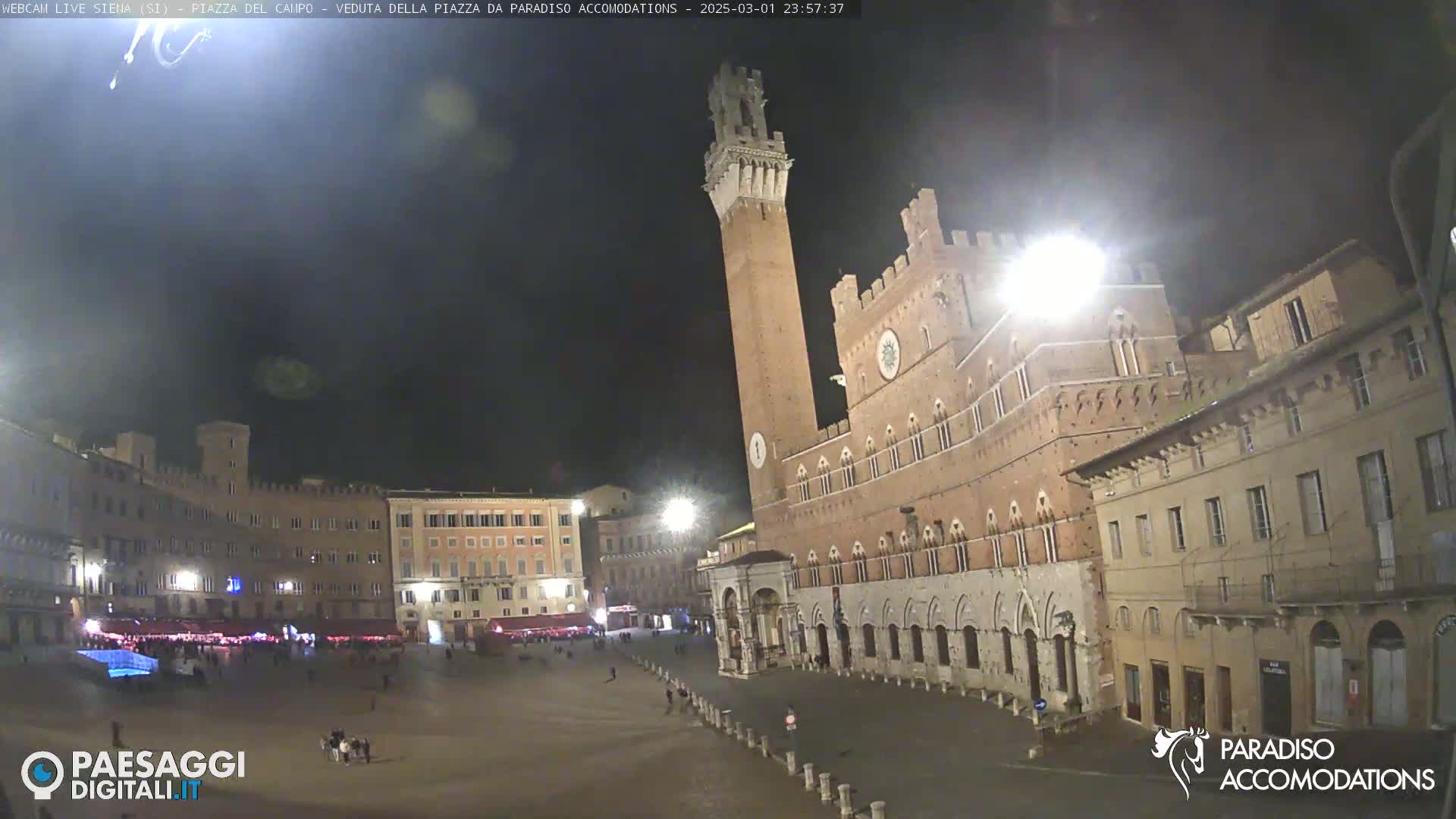 A large, empty plaza at night, surrounded by medieval-style buildings, one of which is a tall tower.