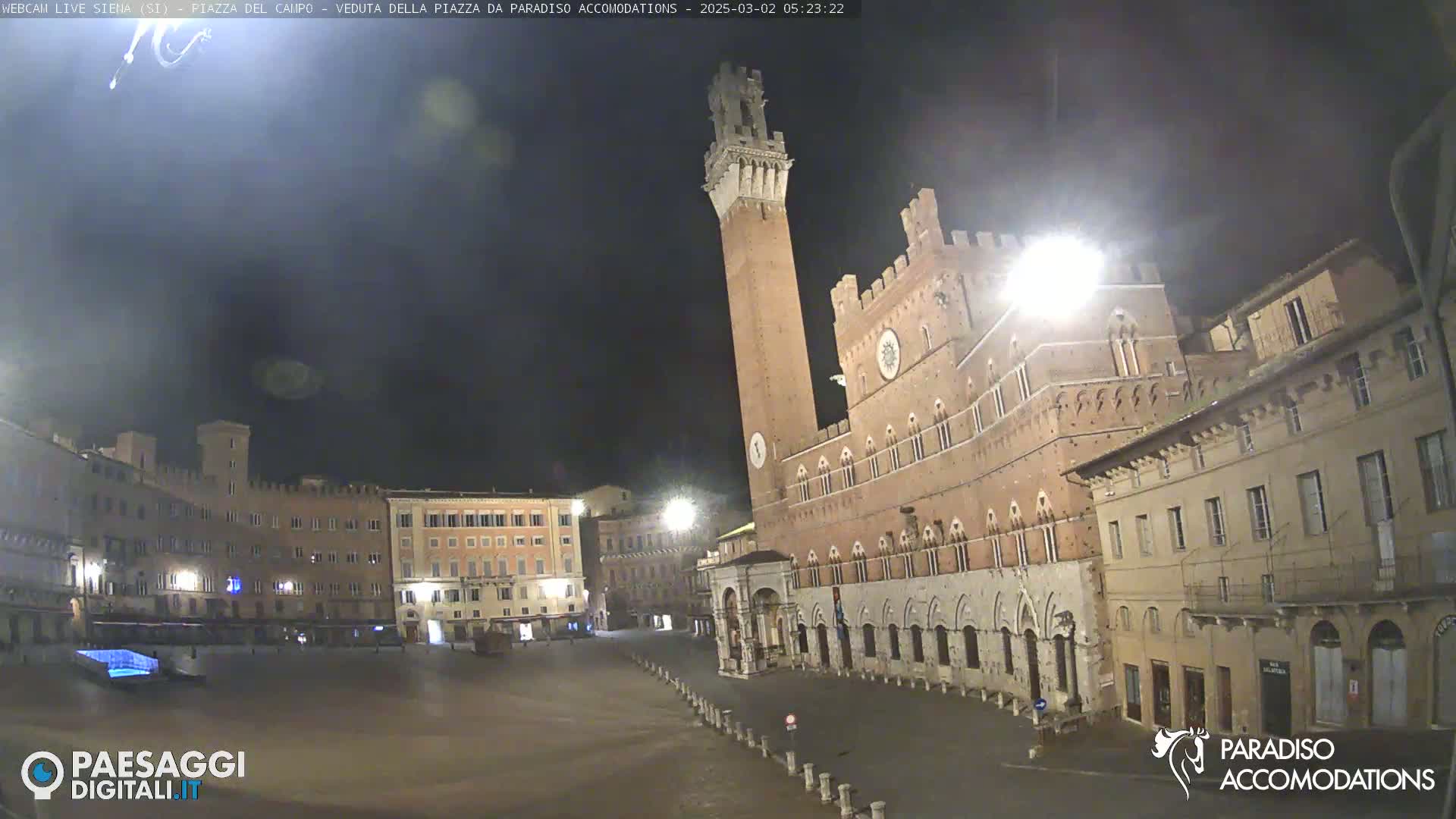 A nighttime view of a large, mostly empty plaza in a European city, featuring a tall brick tower and surrounding historical buildings under a dark sky.