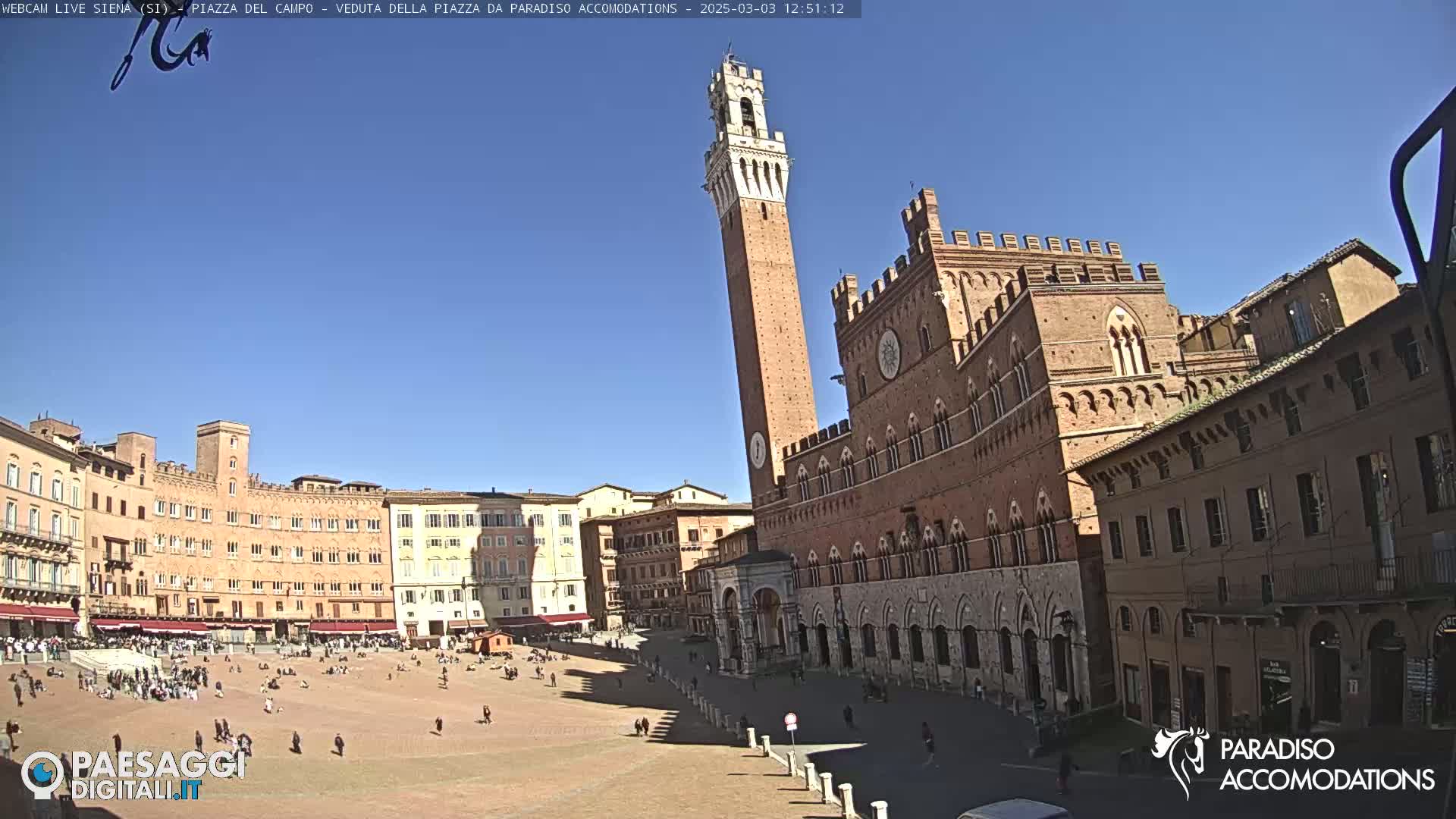 A large, brick piazza in Siena, Italy, is bustling with people under a clear, sunny sky, with a tall tower and medieval buildings surrounding the square.