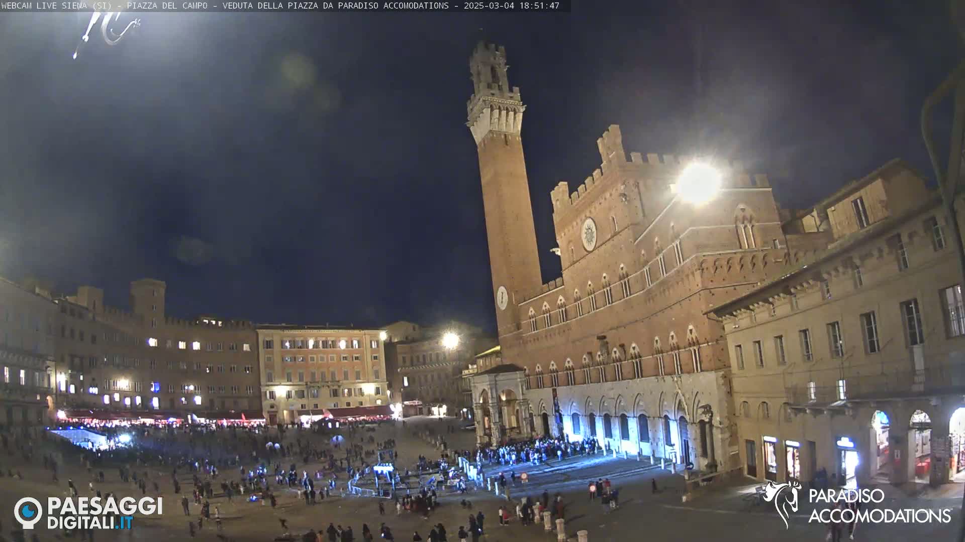 A nighttime view of a large, crowded plaza with a tall brick tower and adjacent buildings under a dark sky.