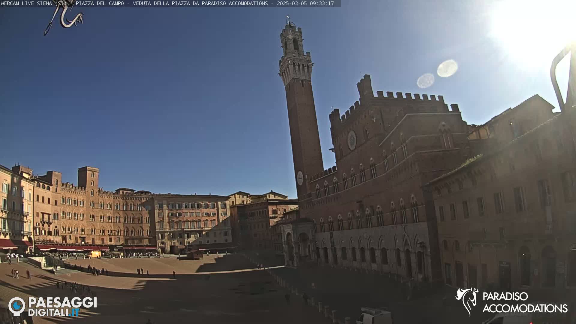 A large, brick public square is shown on a sunny day, with a tall tower and adjacent buildings visible.