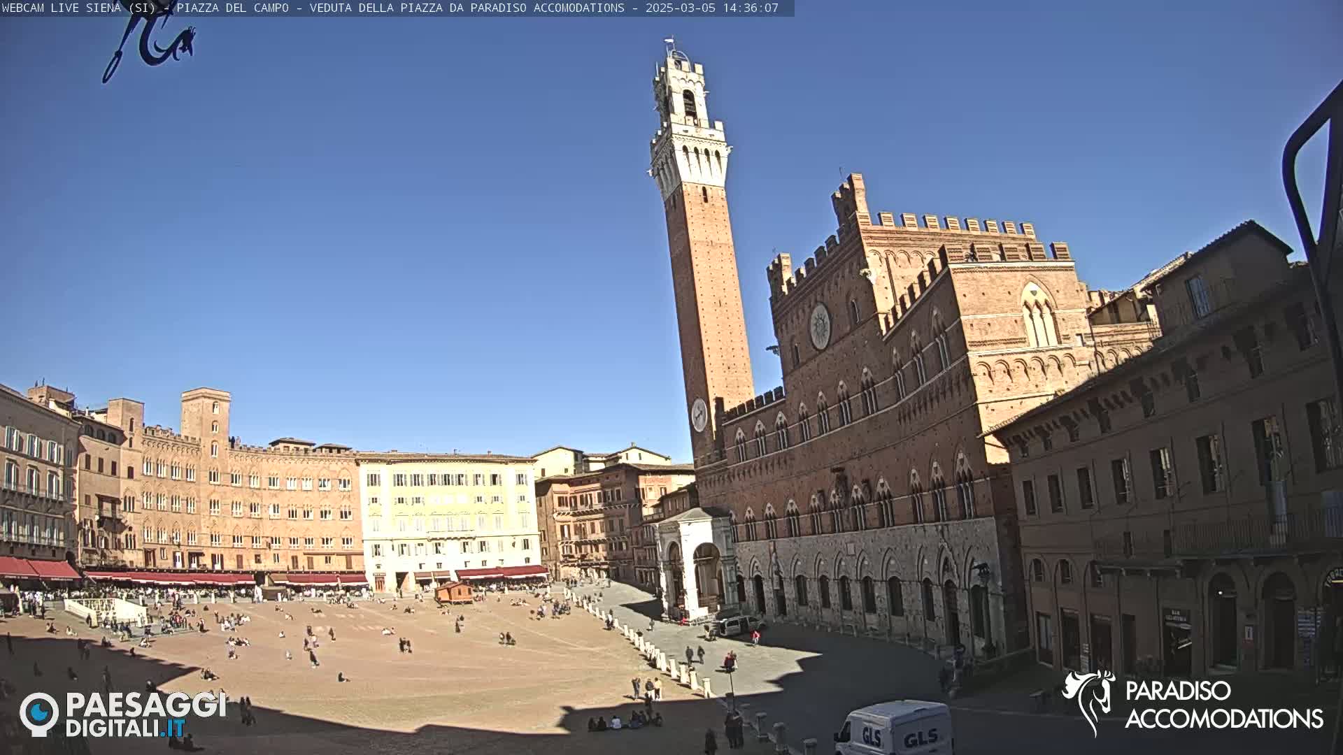 A large, brick public square in Italy is filled with a few people on a sunny day, surrounded by medieval-style buildings, including a tall tower.