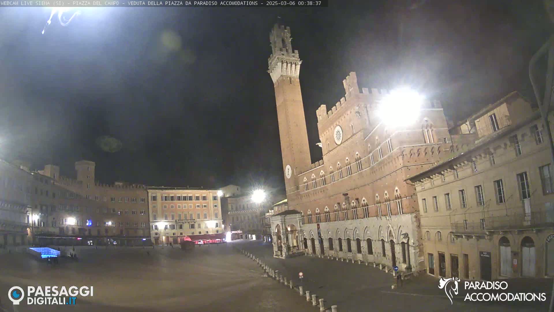 A nighttime view of a large, mostly empty plaza with medieval-style buildings, including a tall tower, under a dark, clear sky.