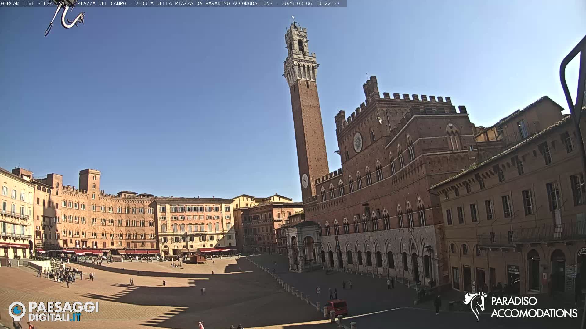 A large, brick public square is shown on a sunny day, featuring a tall tower and several historic buildings.