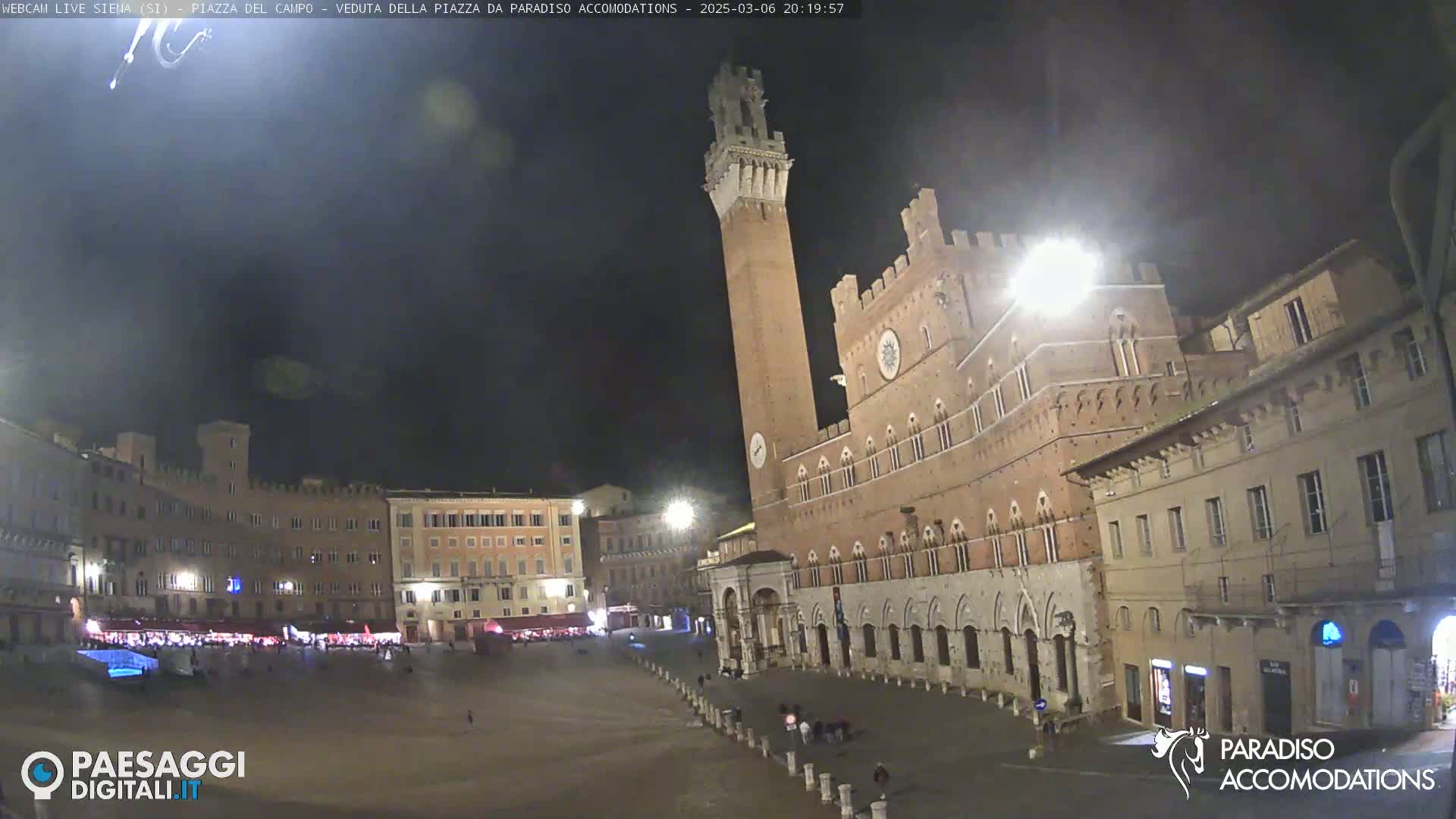 A nighttime view of a large, mostly empty town square in Siena, Italy, featuring a tall brick tower and surrounding medieval-style buildings under a dark, clear sky.