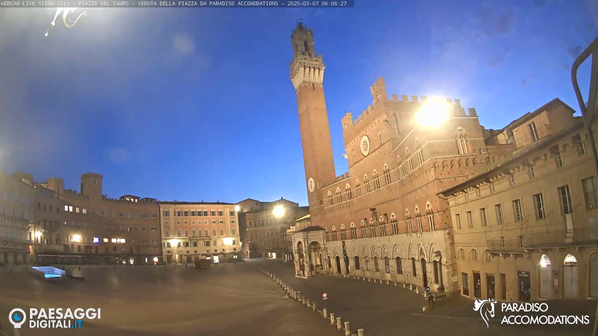 A large, mostly empty town square is shown at dawn under a clear, twilight sky, with a tall brick tower and surrounding medieval buildings.