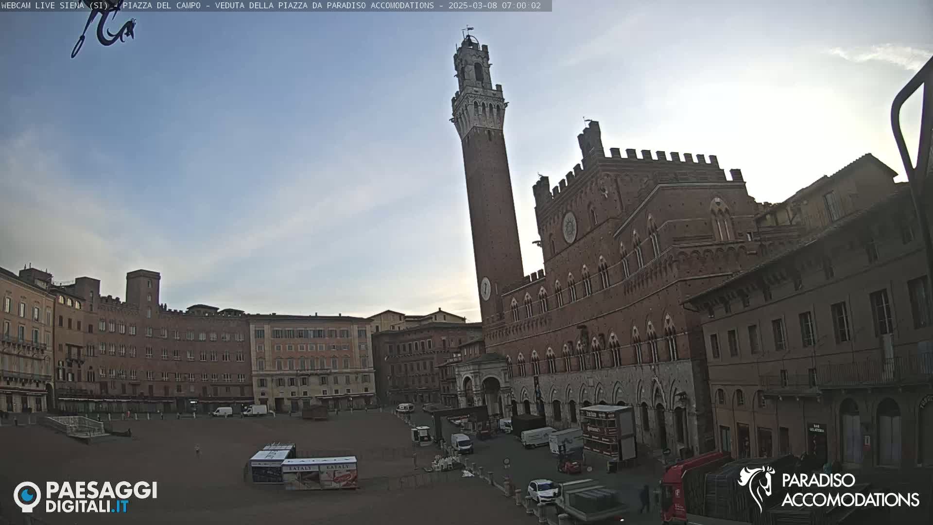 A large, brick public square in the early morning, under a clear pale blue sky, is dominated by a tall, slender tower and a large, ornate brick building.