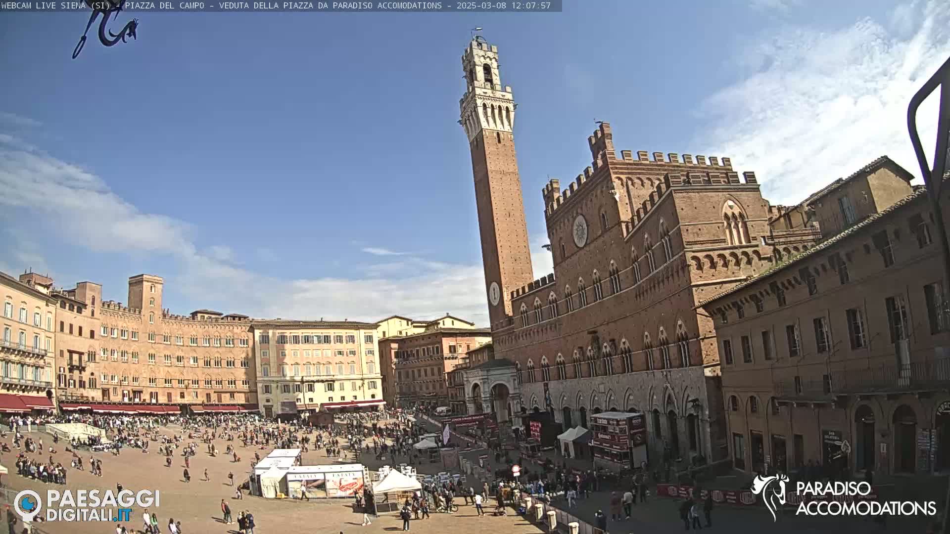 A large, sunny plaza is filled with people, surrounded by historic brick buildings, including a tall tower.