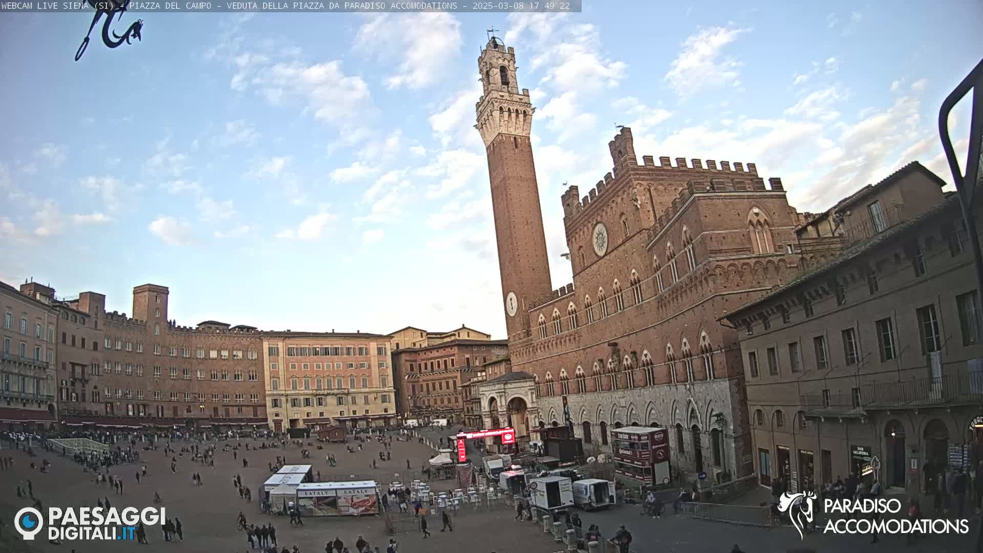 A large, brick public square with a tall tower and several buildings, under a partly cloudy sky.