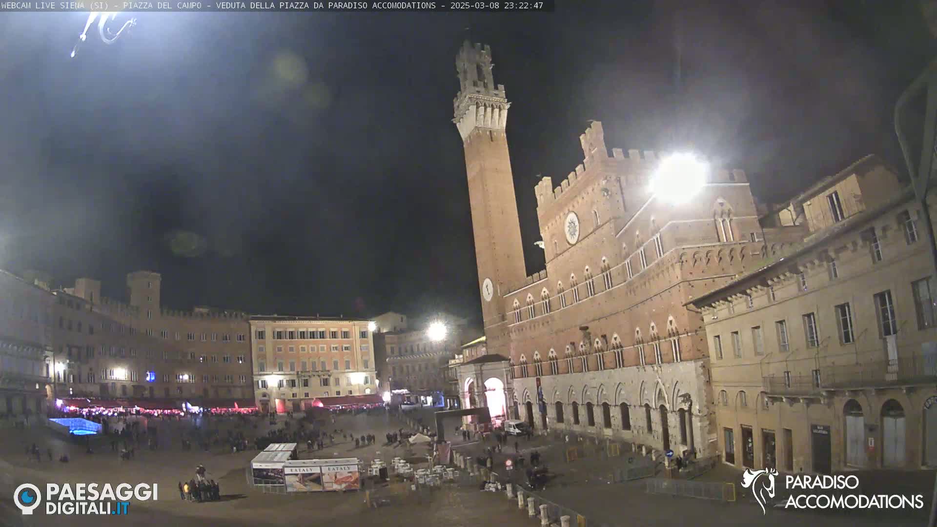 A nighttime view of a large, mostly empty town square with a tall, brick tower and surrounding buildings.