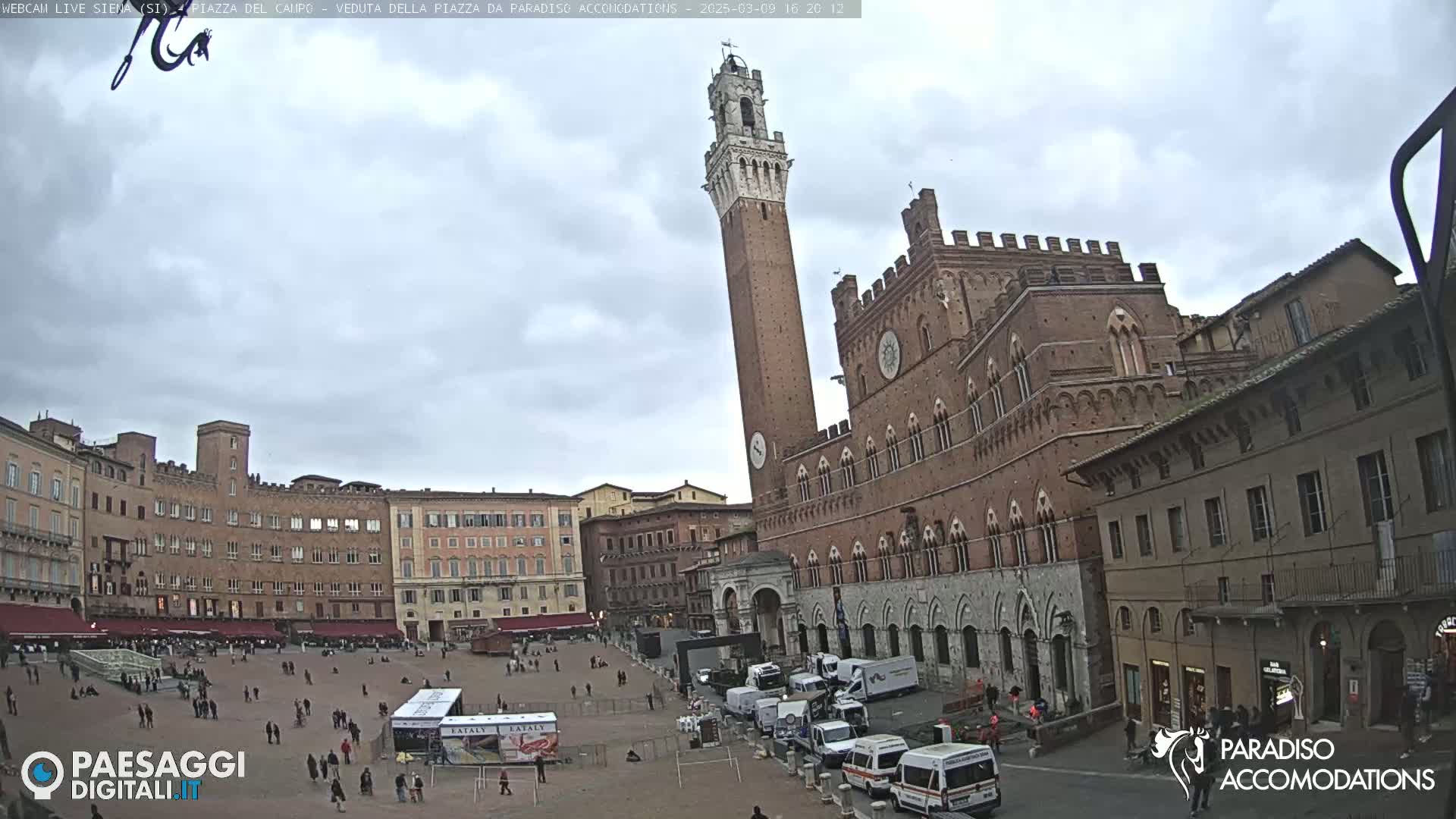 A large, mostly empty square in a historic Italian city, under an overcast sky, is surrounded by brick buildings, including a tall tower and a grand palace.