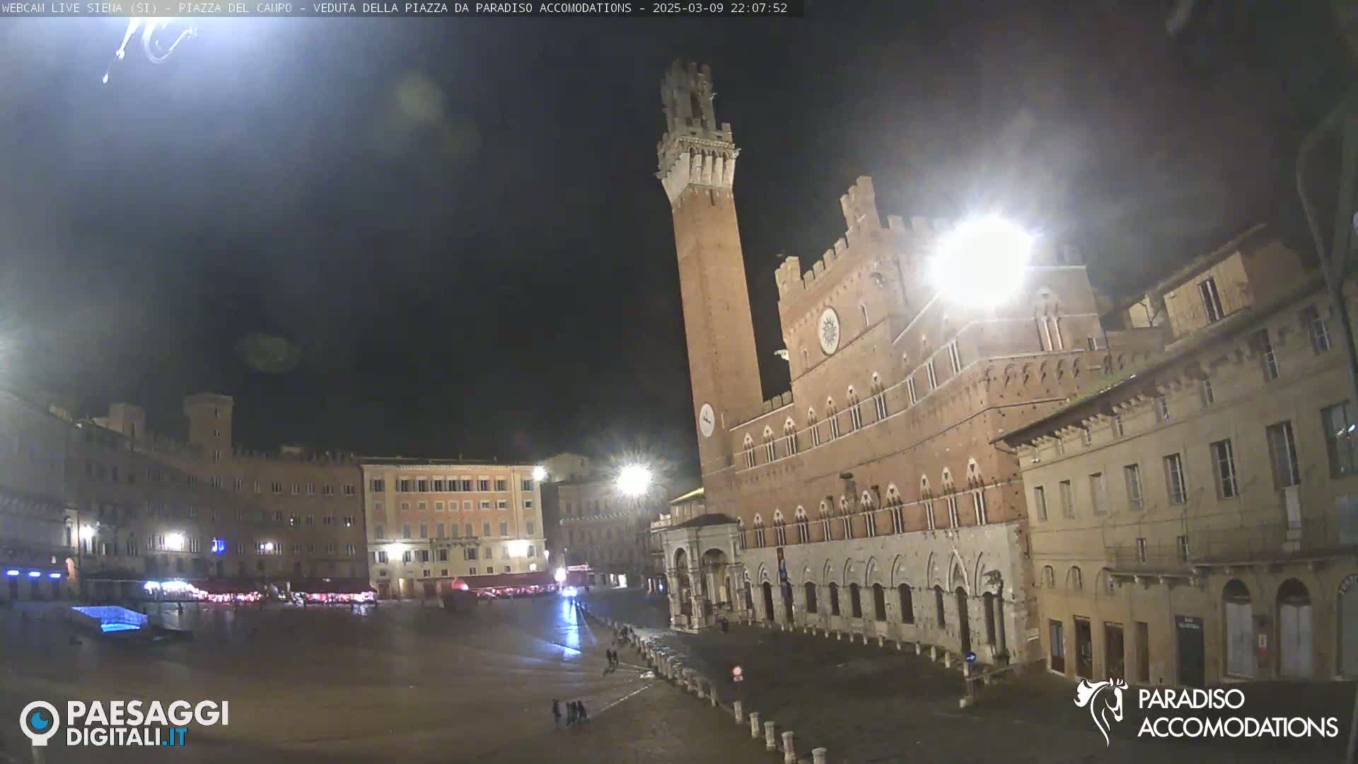 A nighttime view of a large, empty square in a European city with a tall tower and surrounding buildings under a dark sky.