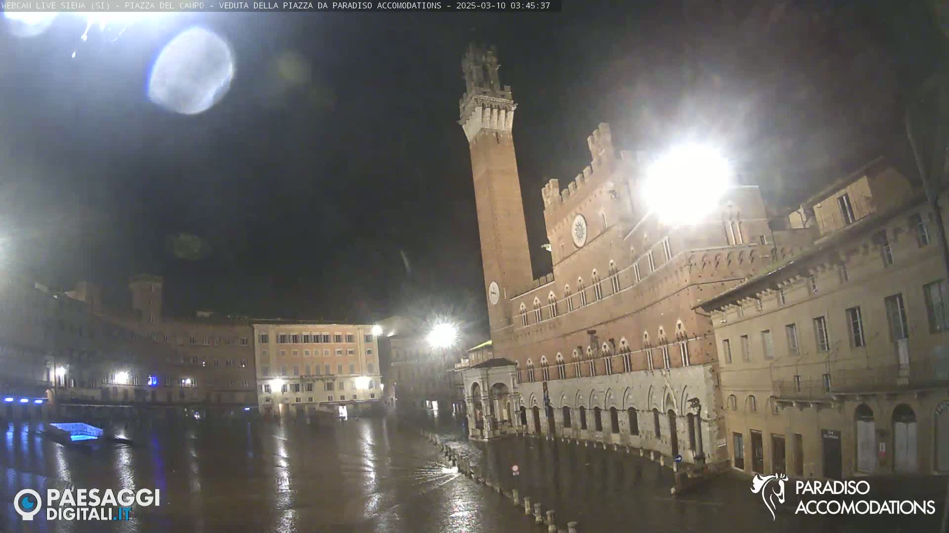 A nighttime view of a flooded town square in the rain, featuring a tall brick tower and surrounding buildings.