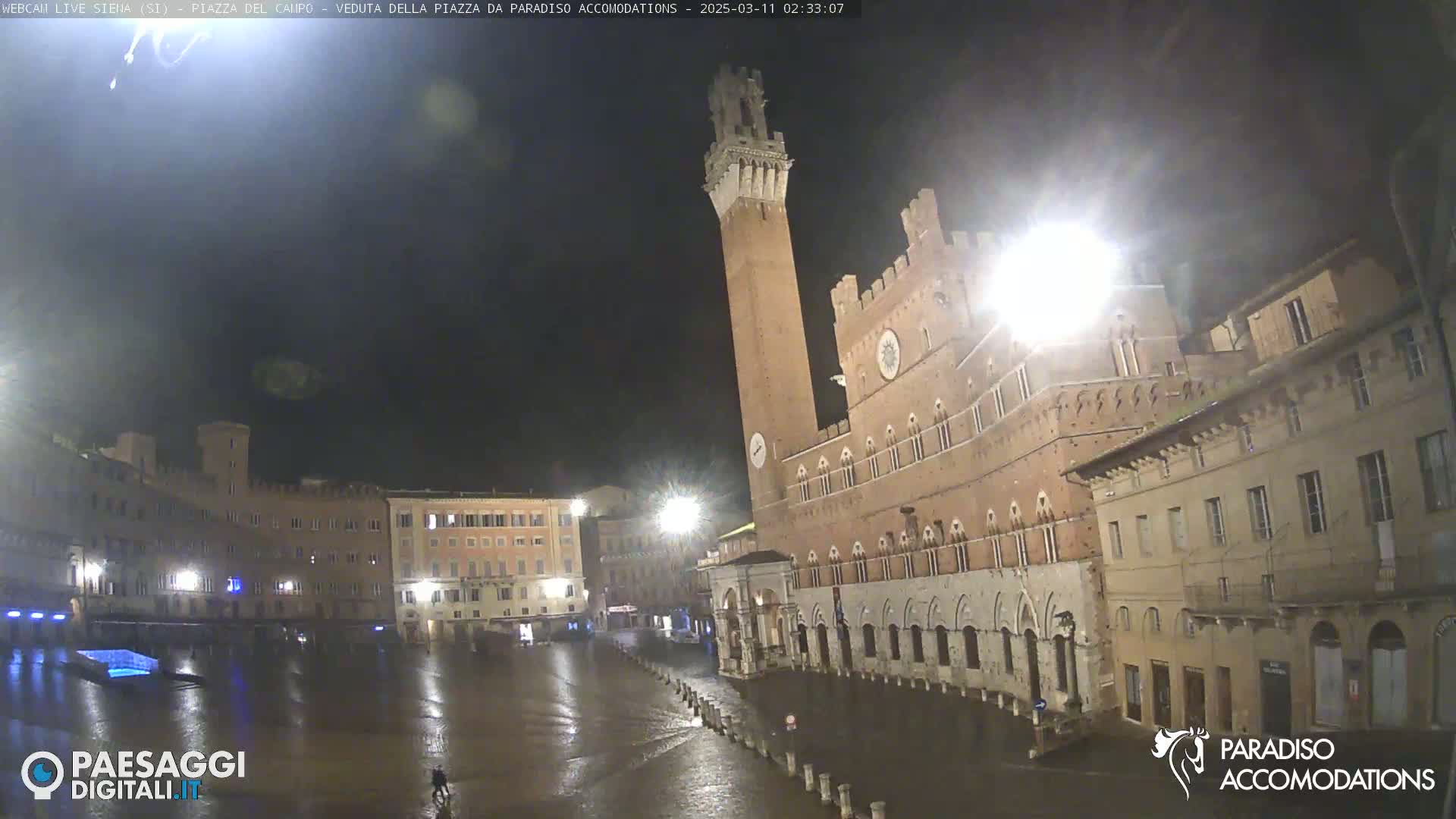 A large, brick plaza at night is wet from rain, with a tall tower and several buildings visible under dim lighting.