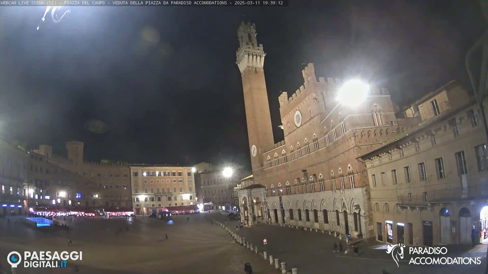 A nighttime view of a large, empty piazza in a city, surrounded by medieval-style buildings, under a clear, dark sky.