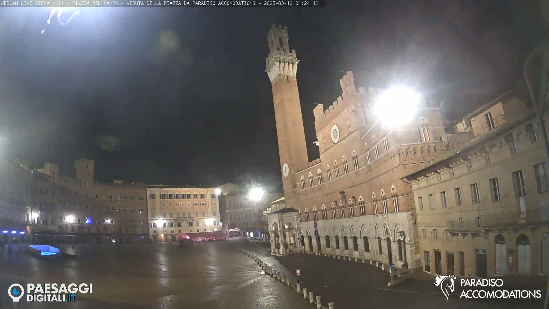 A nighttime view of a large, mostly empty town square in Italy, featuring a tall brick tower and surrounding historic buildings under a dark, clear sky.