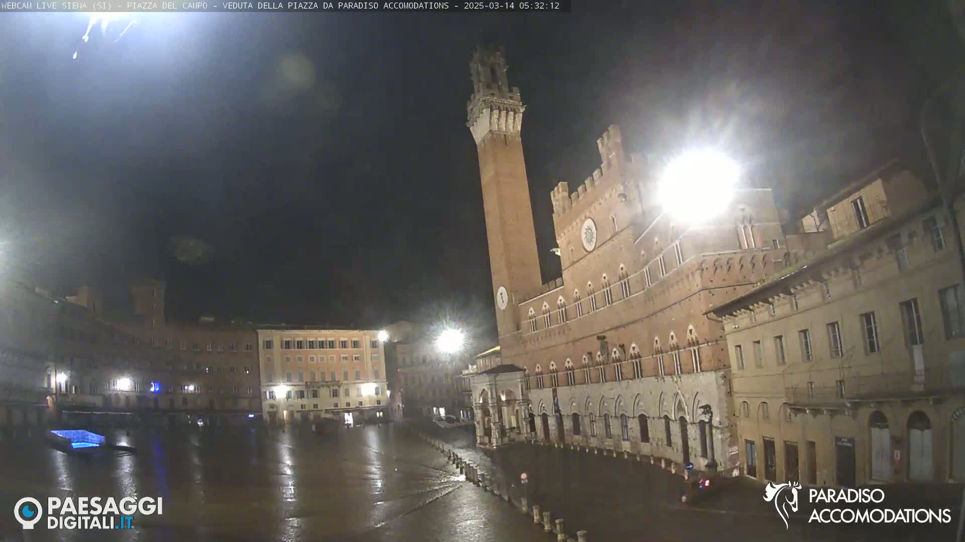 A rain-slicked, nighttime piazza in Italy, featuring a tall brick tower and adjacent medieval-style buildings.