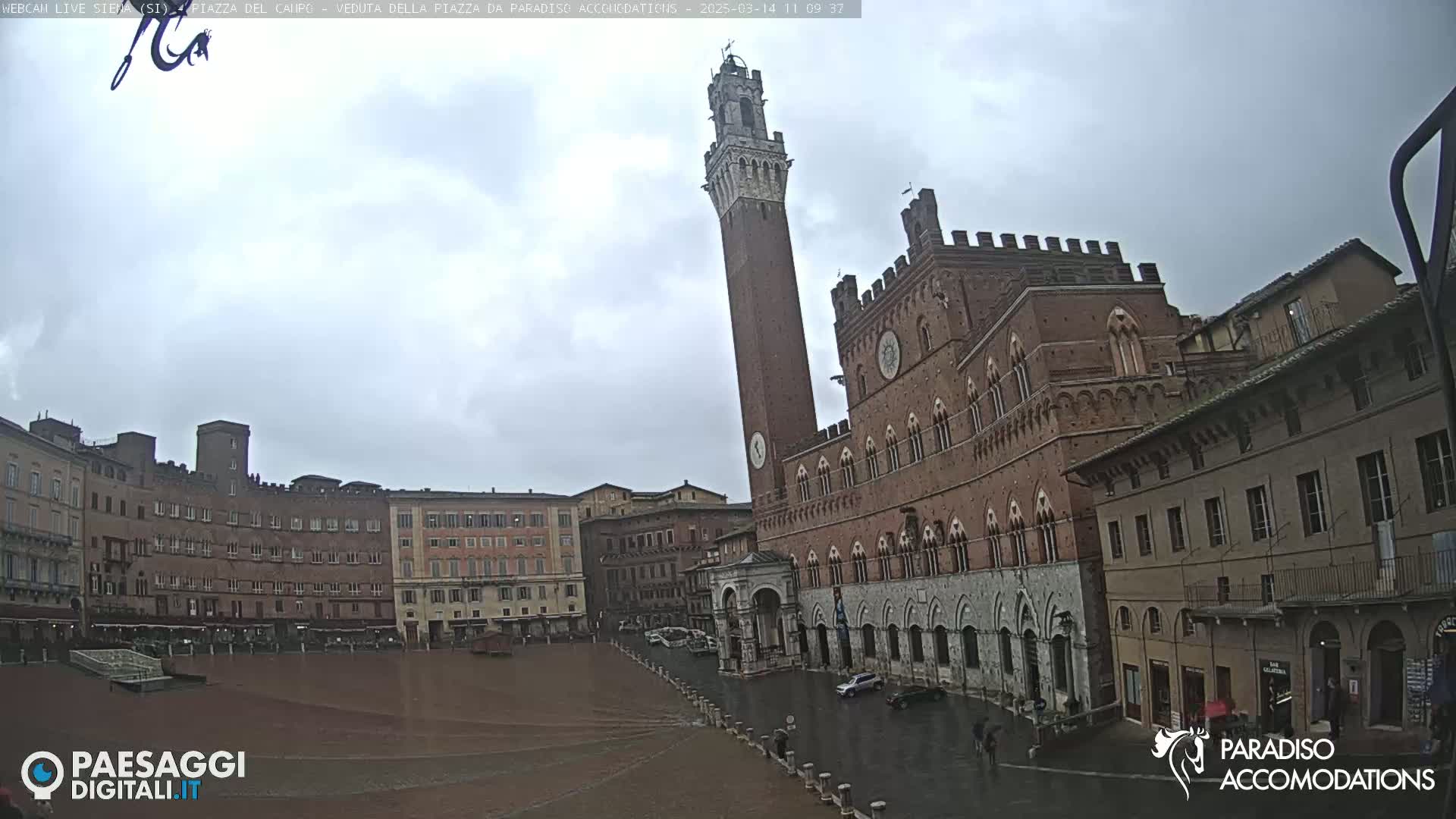 A large brick piazza in Siena, Italy is shown on a rainy day with a tall tower and several buildings surrounding a wide open square.