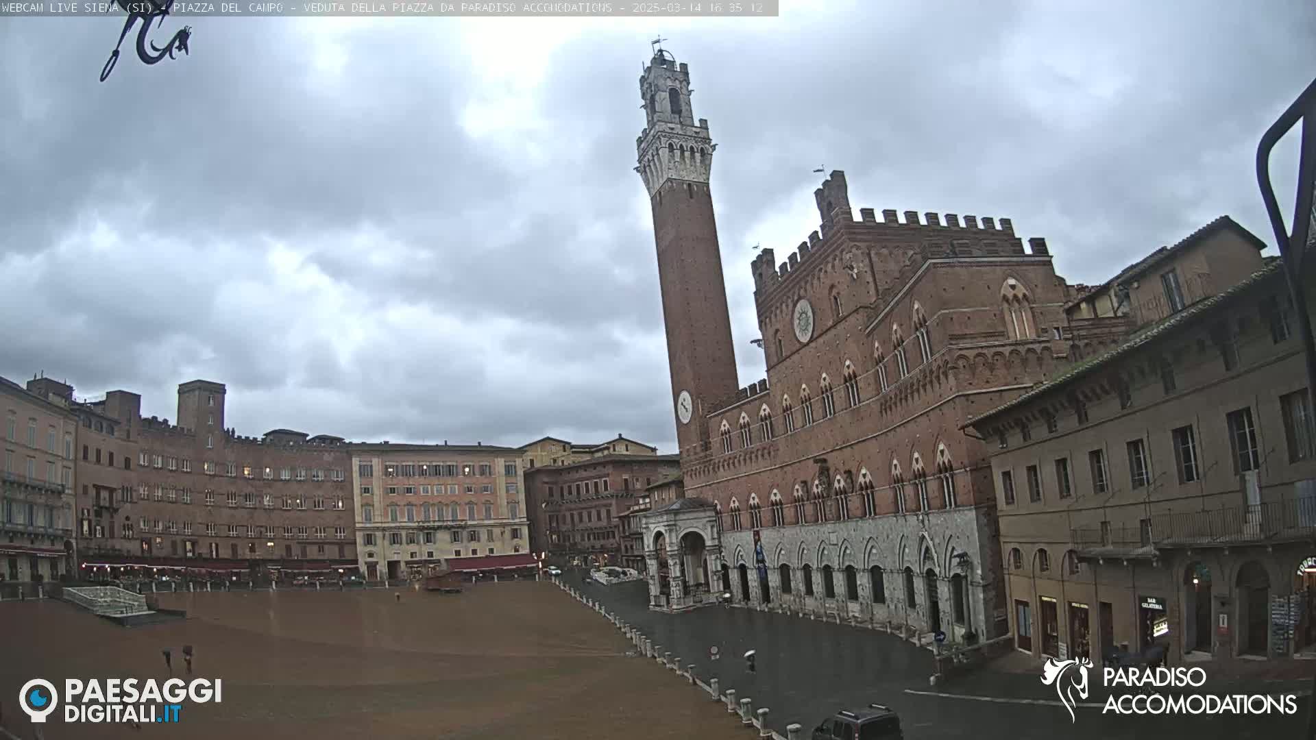 A large, brick piazza in Italy is shown on an overcast day with a tall tower and several buildings surrounding the open square.