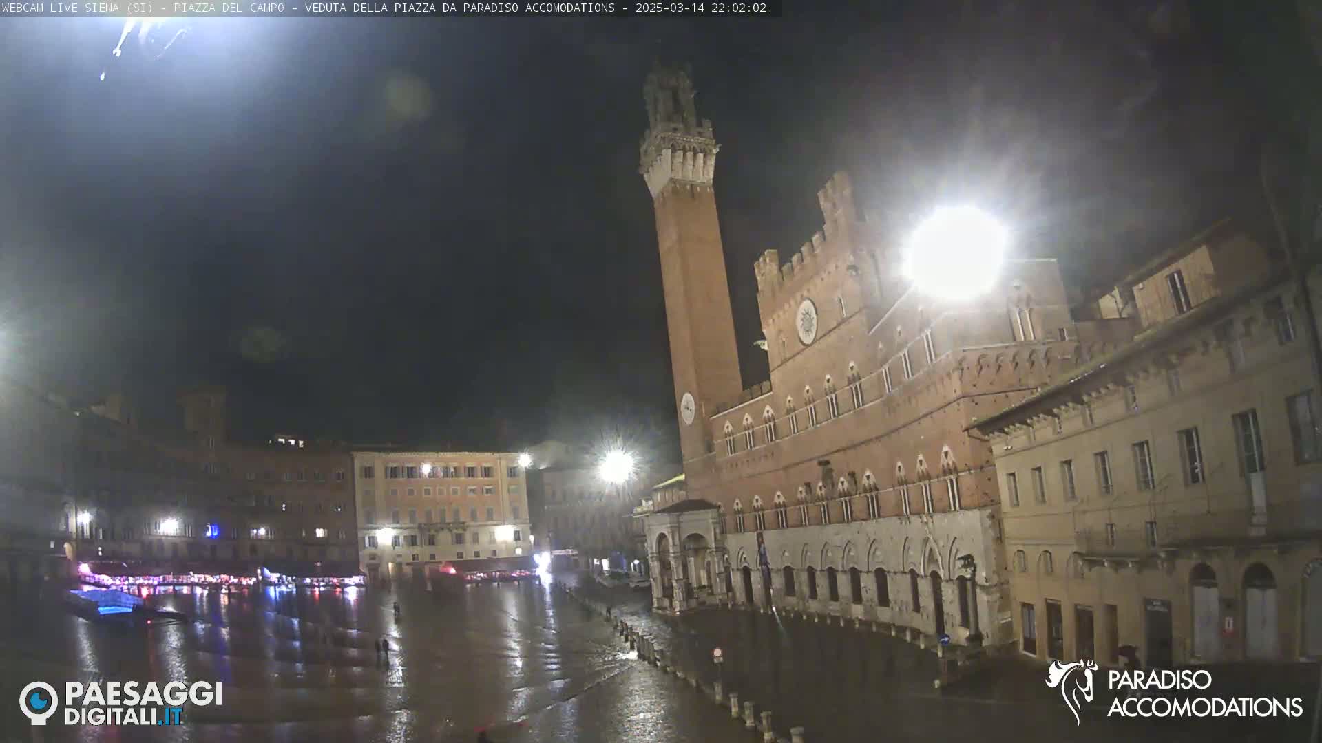 A nighttime view of a flooded plaza in a European city, featuring a tall brick tower and several historic buildings under dark skies.
